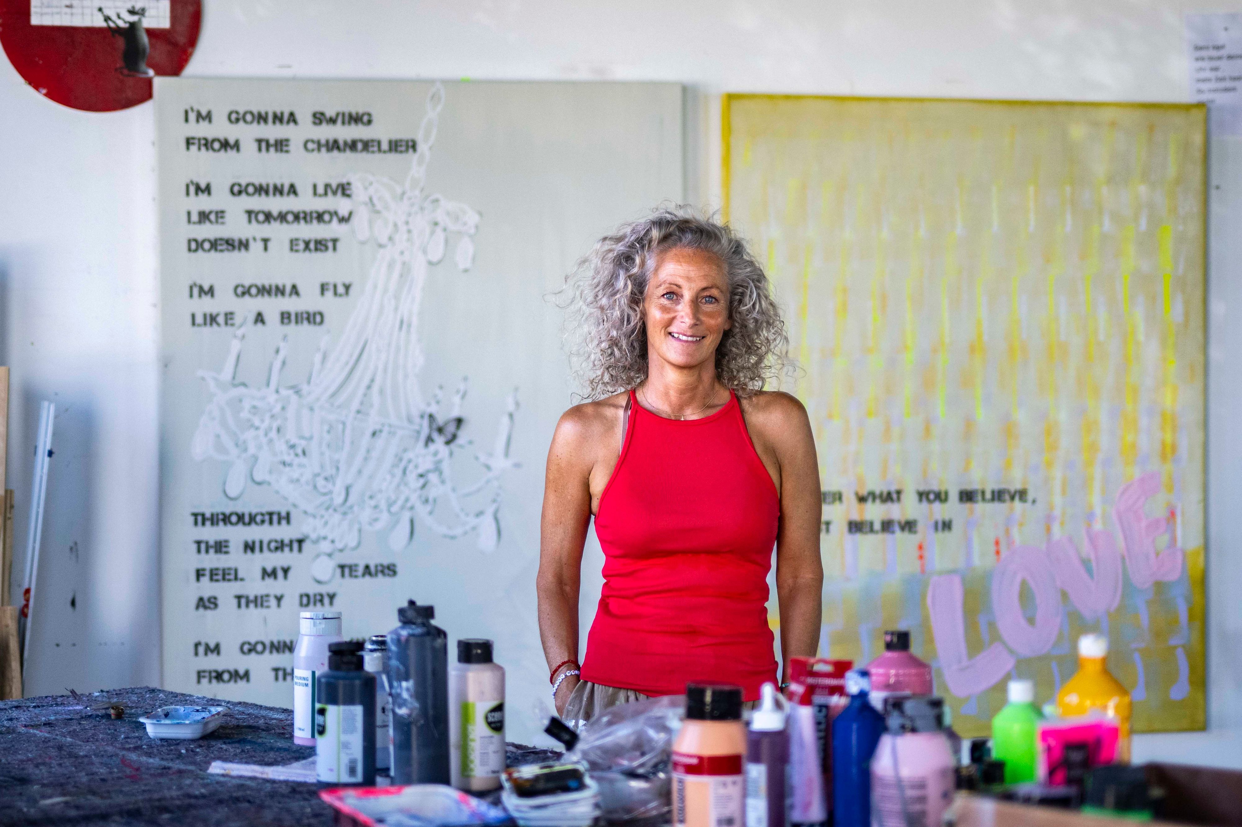 Eine Frau mit lockigen Haaren steht an einem Tisch mit Farben und Sprühdosen in einem Atelier.