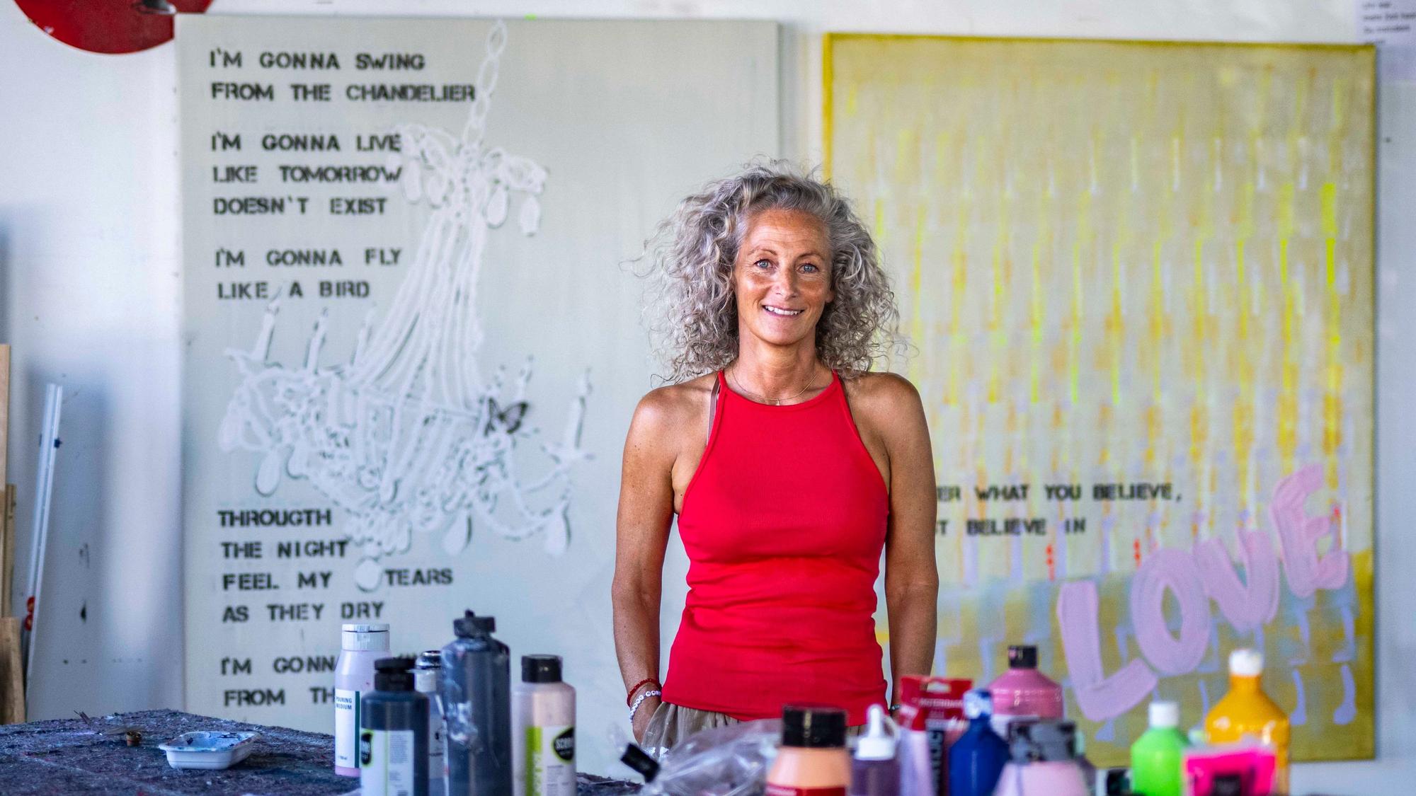 Eine Frau mit lockigen Haaren steht an einem Tisch mit Farben und Sprühdosen in einem Atelier.