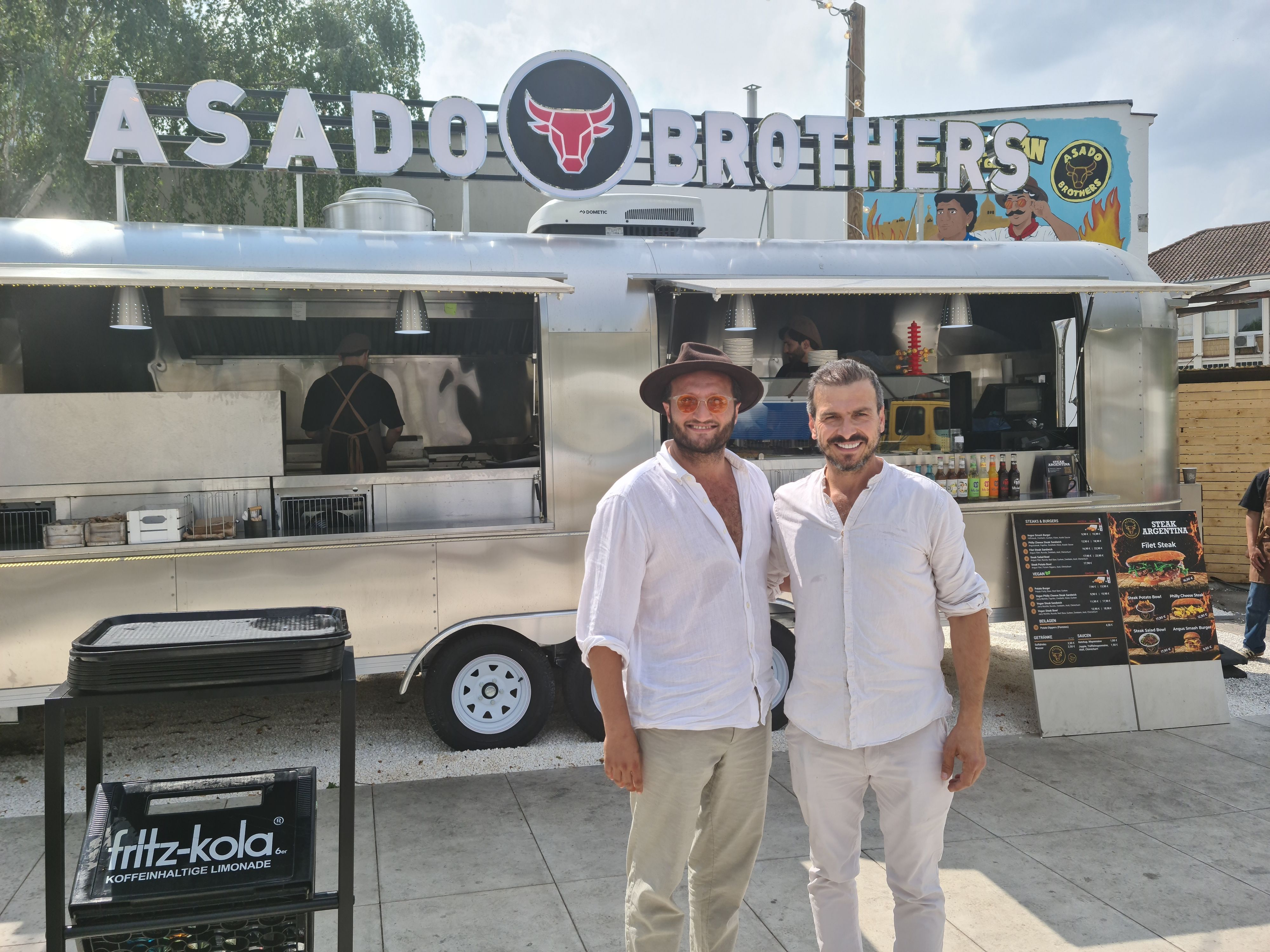 Zwei Männer in weißen Leinenhemden stehen vor einem silbernen Foodtruck.