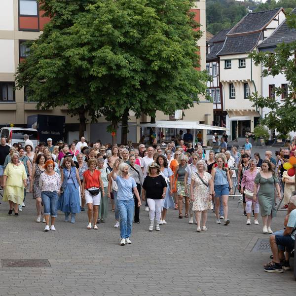 Viele Tanzbegeisterte, vornehmlich Frauen, stehen hinter der Tanzlehrerin, die ihren rechten Arm erhoben hat.