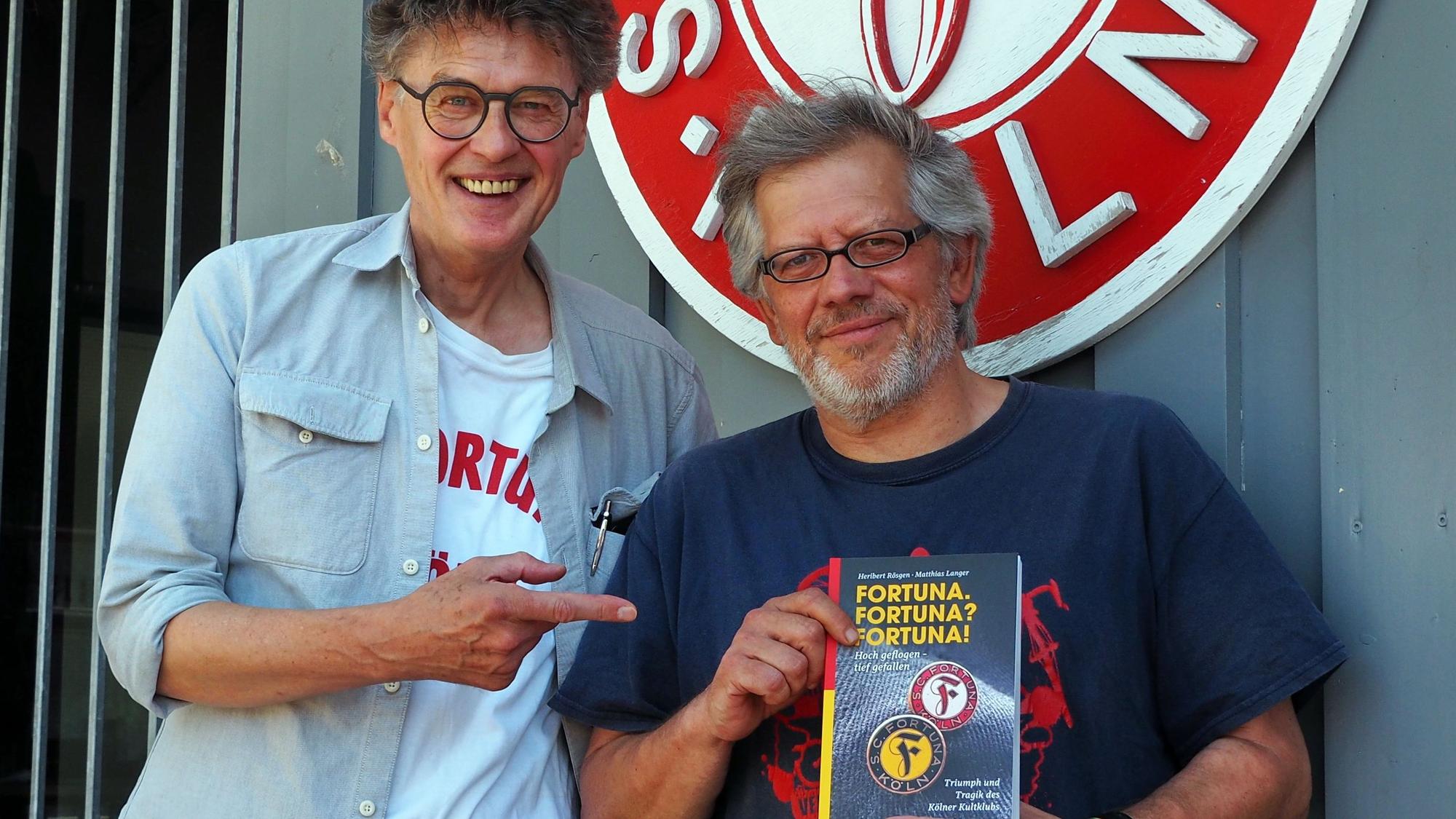 Zwei Männer stehen vor dem Vereinslogo von Fortuna Köln, der Mann rechts hat das neue Buch in der Hand.