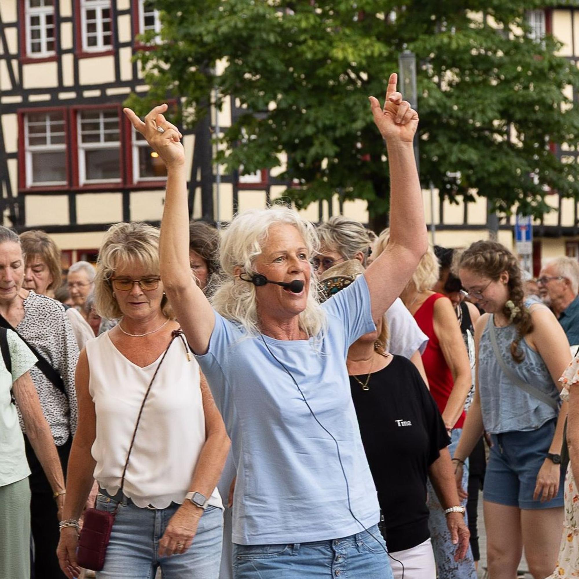 Die Tanzlehrerin hat beide Arme erhoben. Dahinter stehen mehrere Frauen.