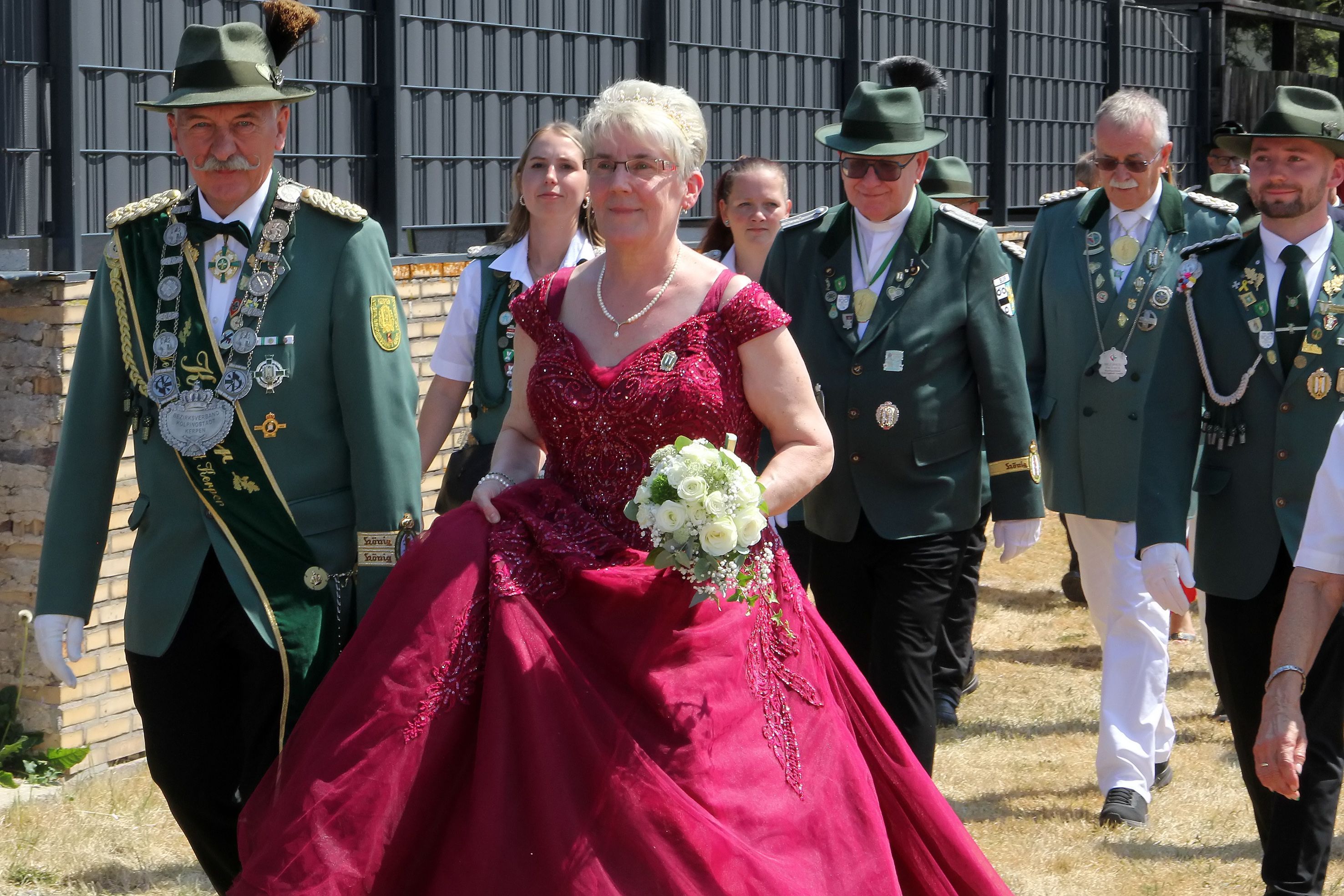 Männer und Frauen in Schützenuniformen und eine Frau in rotem Kleid gehen durch einen Ort.