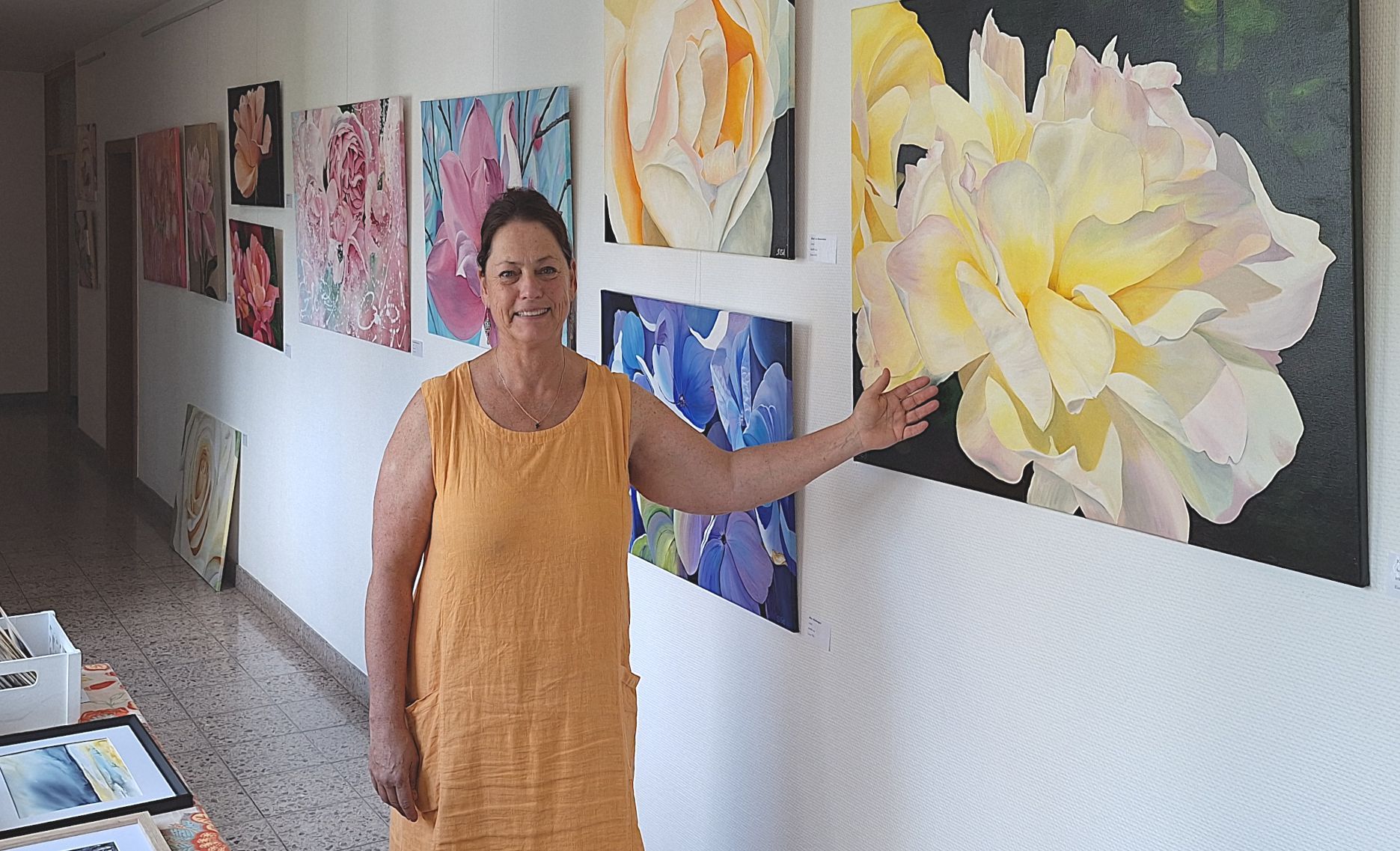 Eine Frau in eine gelben Kleid zeigt auf Bilder einer Ausstellung, die Blüten zeigen.