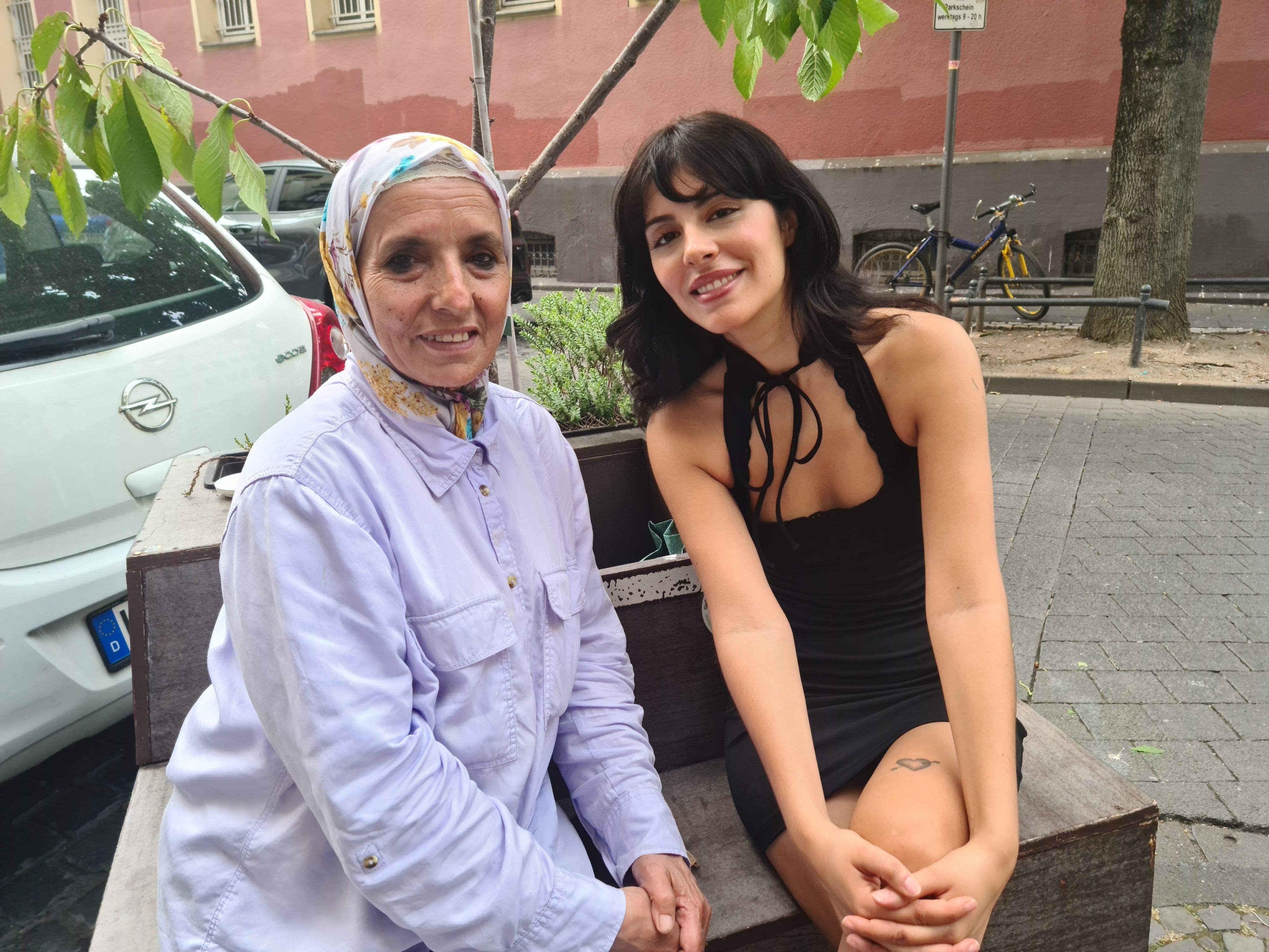 Eine Frau mit Kopftuch und im helllila Hemd sitzt neben einer jungen Frau im schwarzen Kleid auf der Straße auf einer mobilen Baum-Beet-Bank.