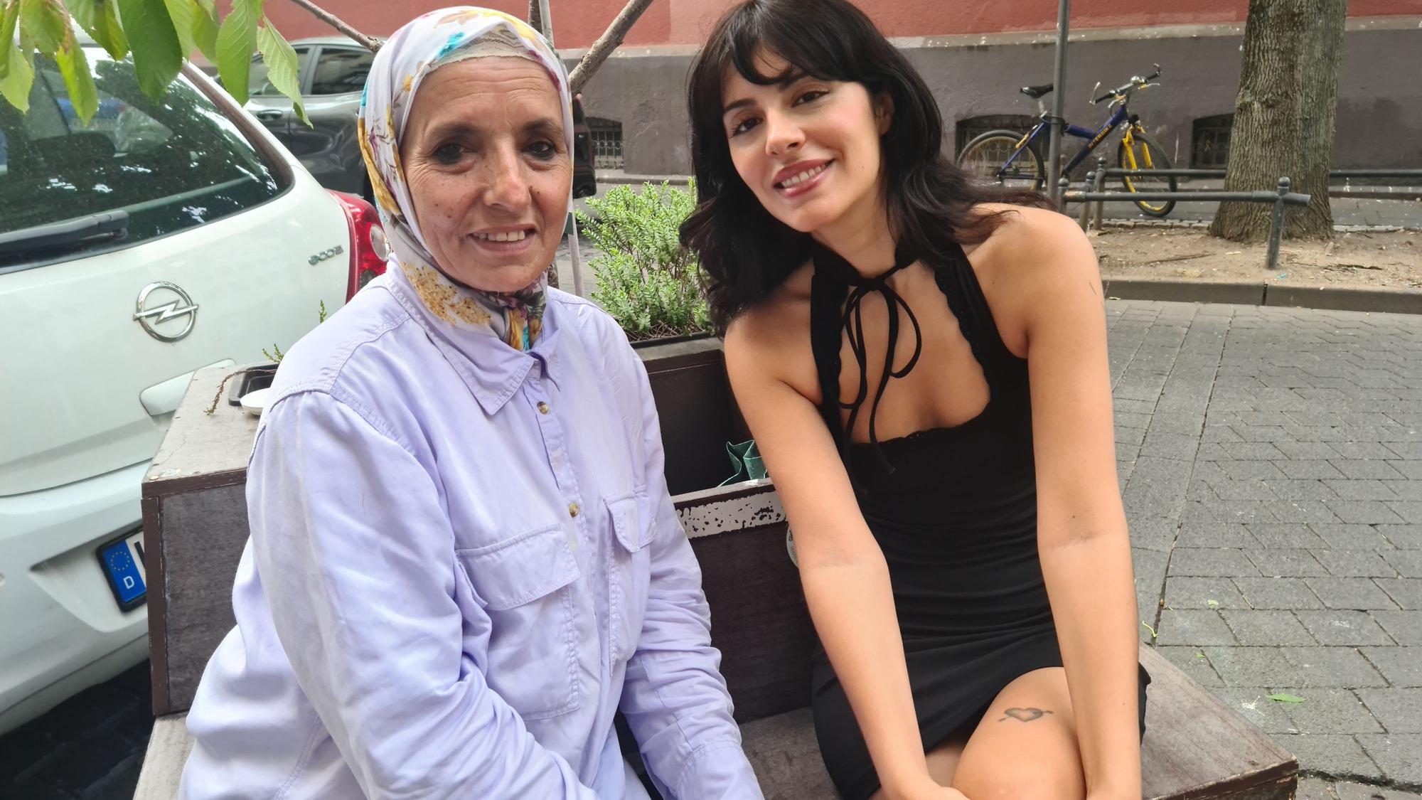Eine Frau mit Kopftuch und im helllila Hemd sitzt neben einer jungen Frau im schwarzen Kleid auf der Straße auf einer mobilen Baum-Beet-Bank.