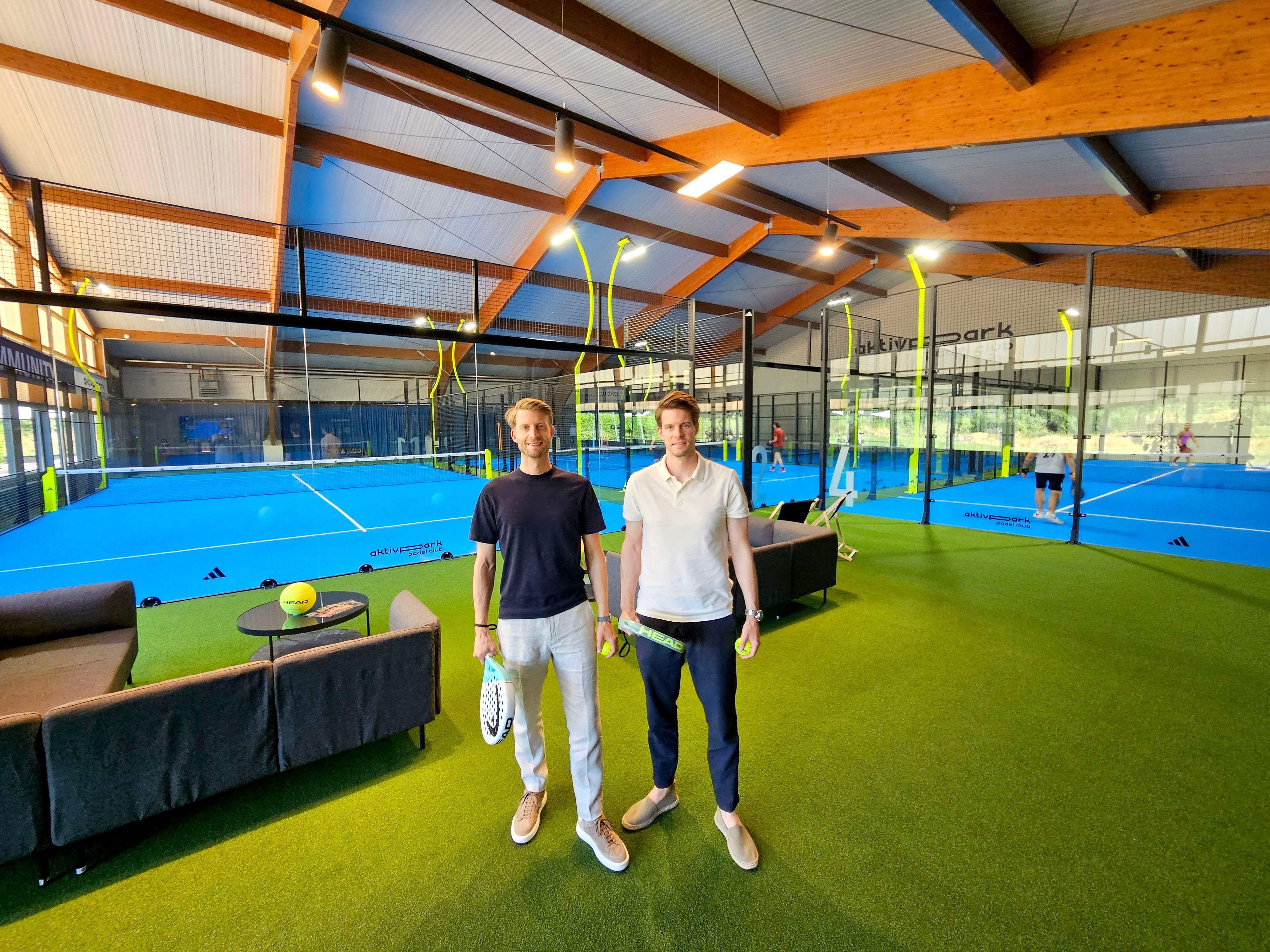 Die beiden Männer mit Padel-Schlägern stehen im Vordergrund, im Hintergrund sind Padel-Plätze zu sehen.