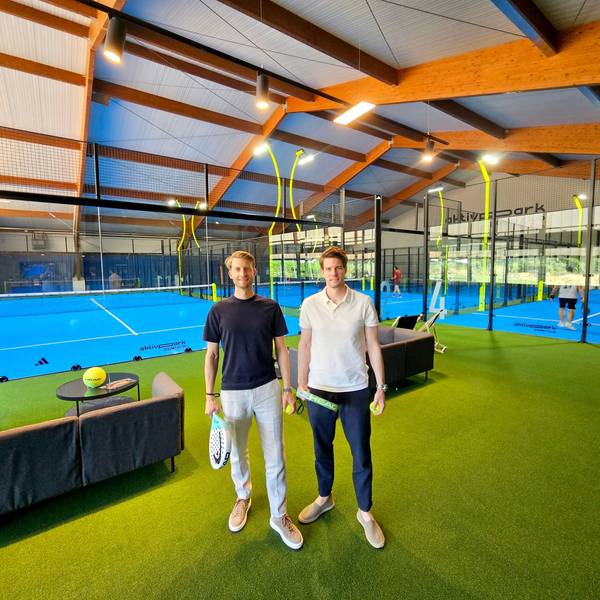 Die beiden Männer mit Padel-Schlägern stehen im Vordergrund, im Hintergrund sind Padel-Plätze zu sehen.