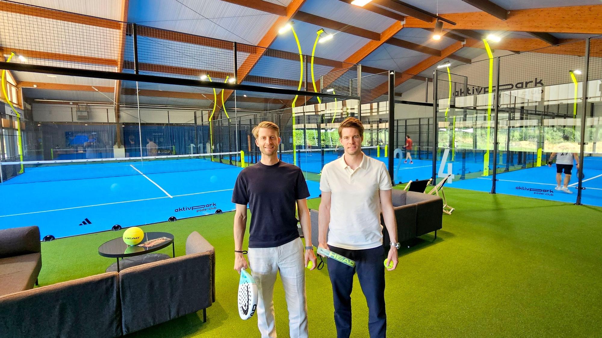 Die beiden Männer mit Padel-Schlägern stehen im Vordergrund, im Hintergrund sind Padel-Plätze zu sehen.