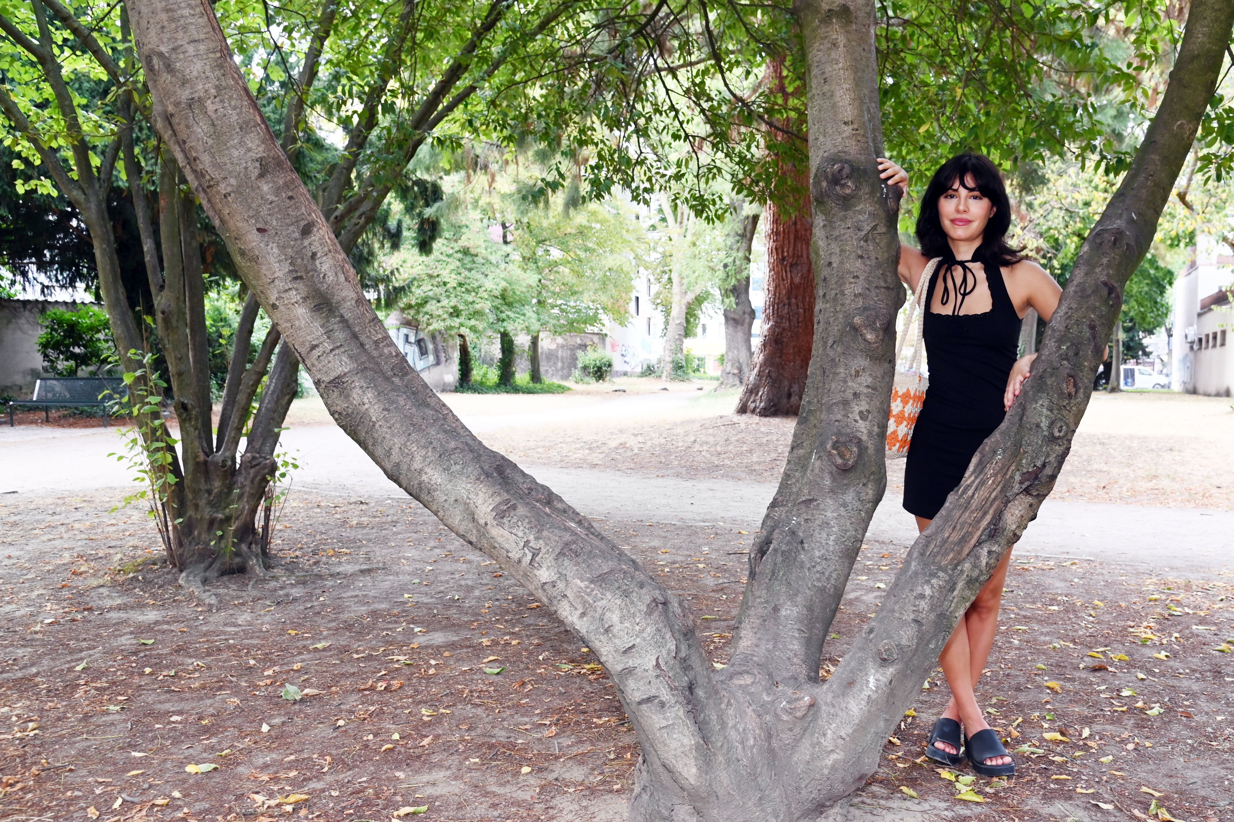 Eine Frau im schwarzen Kleid steht hinter einem Baum, der drei Stämme hat.