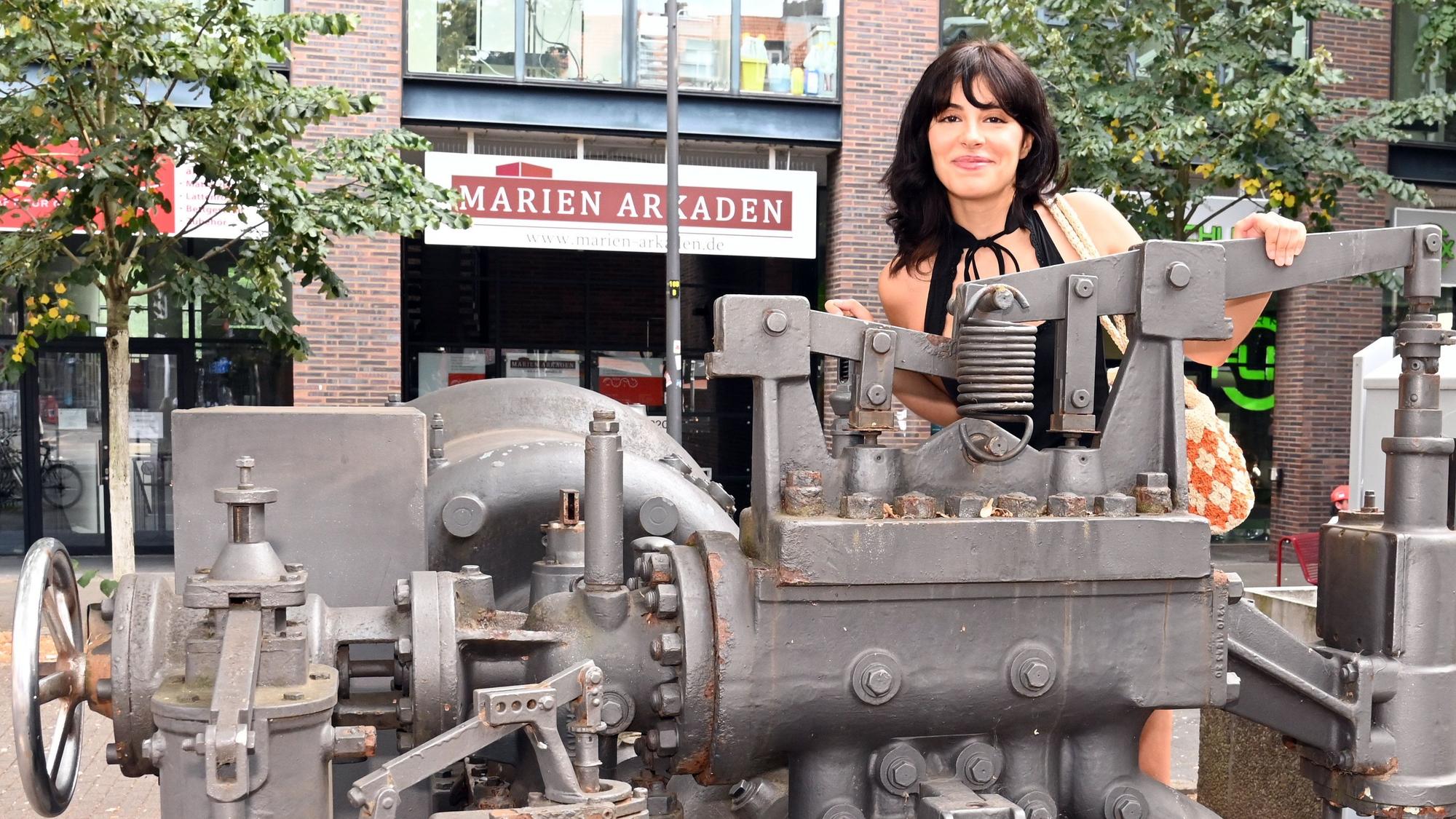 Eine junge Frau mit schwarzem Haar lugt über eine Industriemaschine, die als Denkmal an der Kalker Hauptstraße steht.