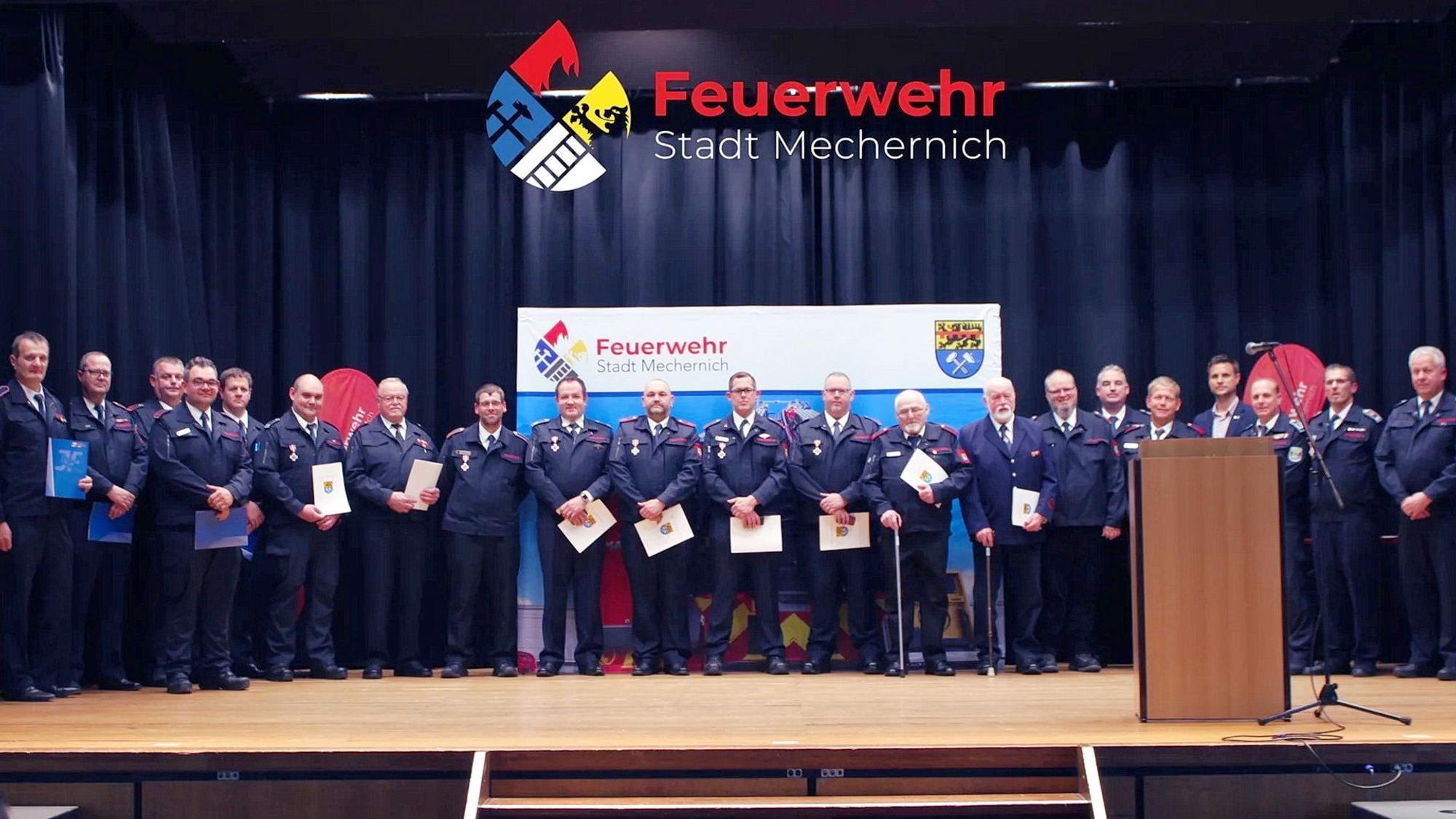 Zahlreiche Feuerwehrangehörige stehen in ihrer Ausgehuniform auf der Bühne der Mechernicher Schulaula. Einige halten Urkunden in der Hand.