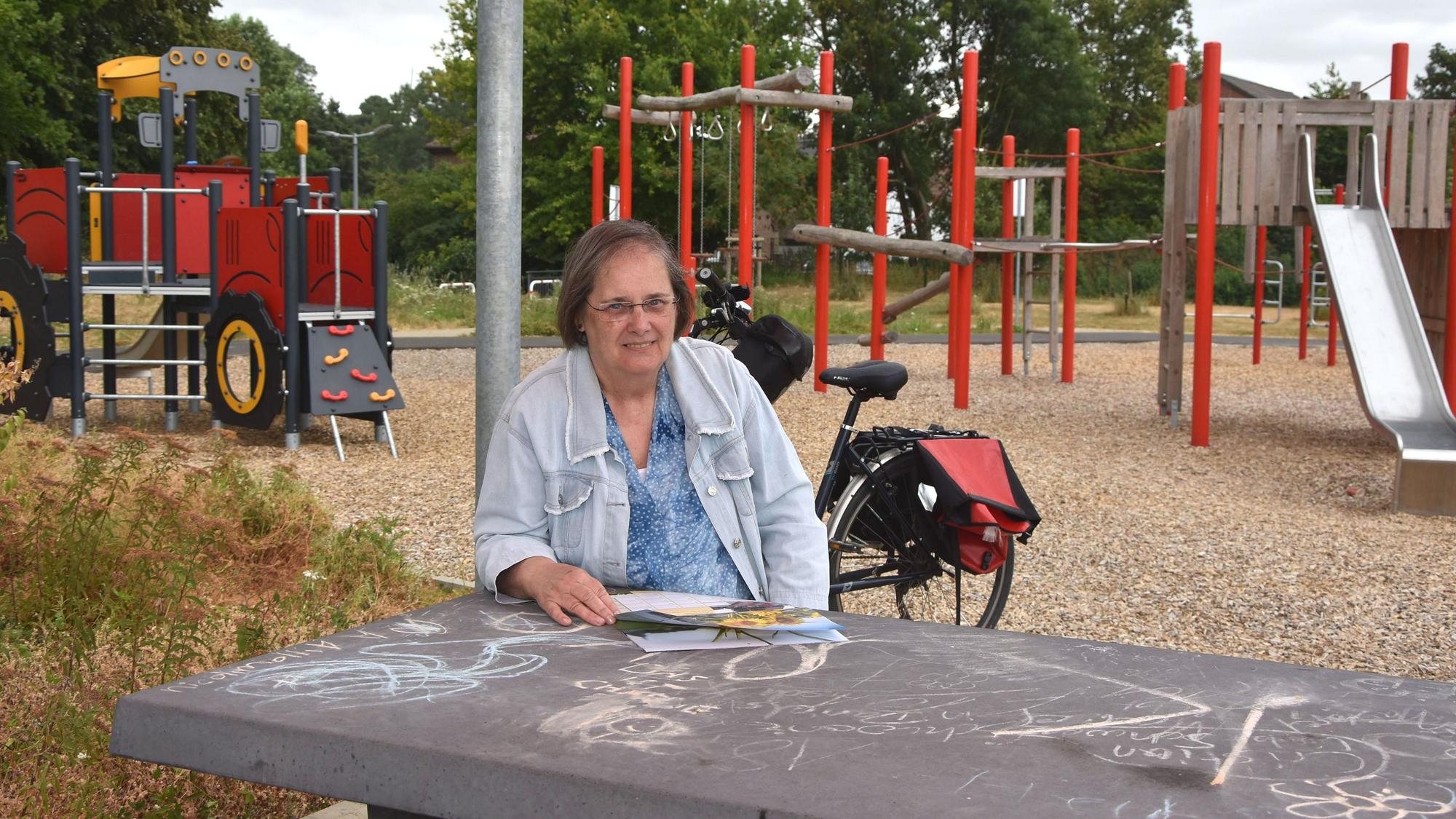 Das Foto zeigt eine Frau, die an einem Tisch vor einem Spielplatz sitzt.