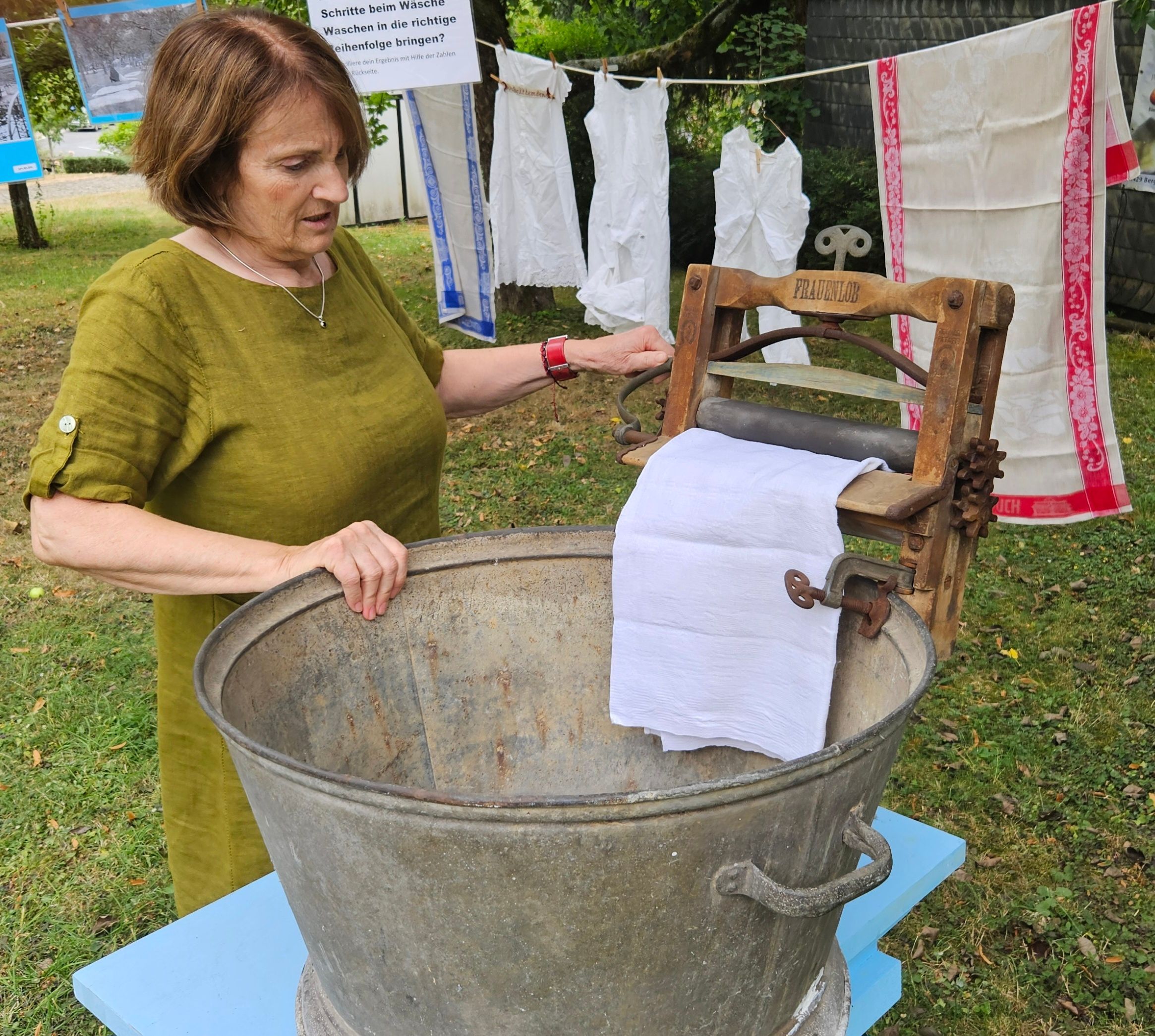 Eine Frau steht an einem Bottich mit einer historischen Wringe, die das Wasser aus einem Wäschestück walzt.