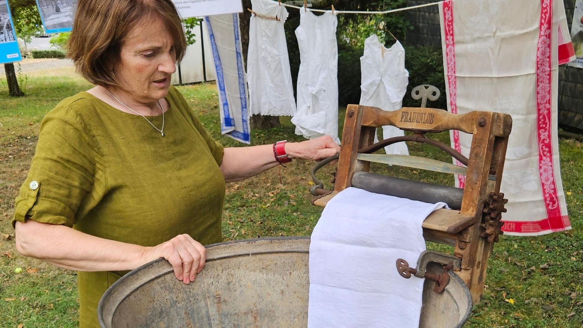 Eine Frau steht an einem Bottich mit einer historischen Wringe, die das Wasser aus einem Wäschestück walzt.