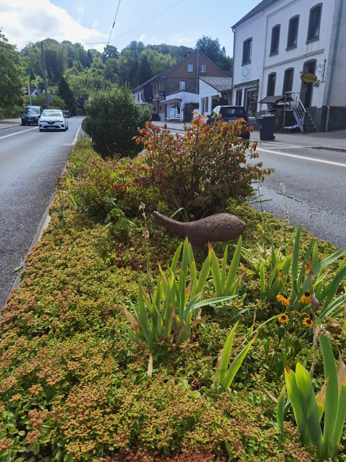 Zu sehen ist eine bepflanzte Verkehrsinsel mit einem Deko-Fisch aus Sandstein.