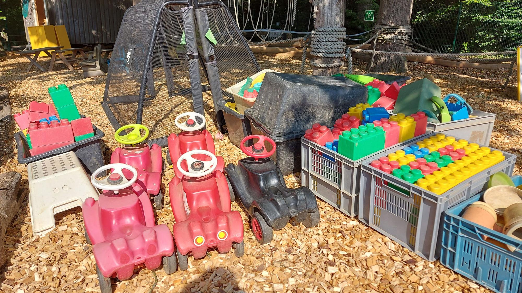 Fünf bunte Bobbycars und große Legosteine aus Kunststoff stehen auf groben Holzspänen.
