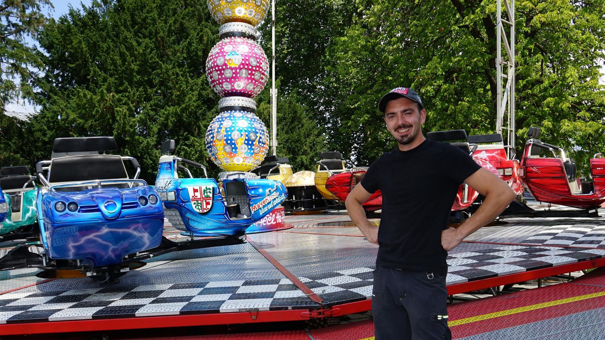 Ein junger Mann steht lächelnd vor einem Kirmes Fahrgeschäft