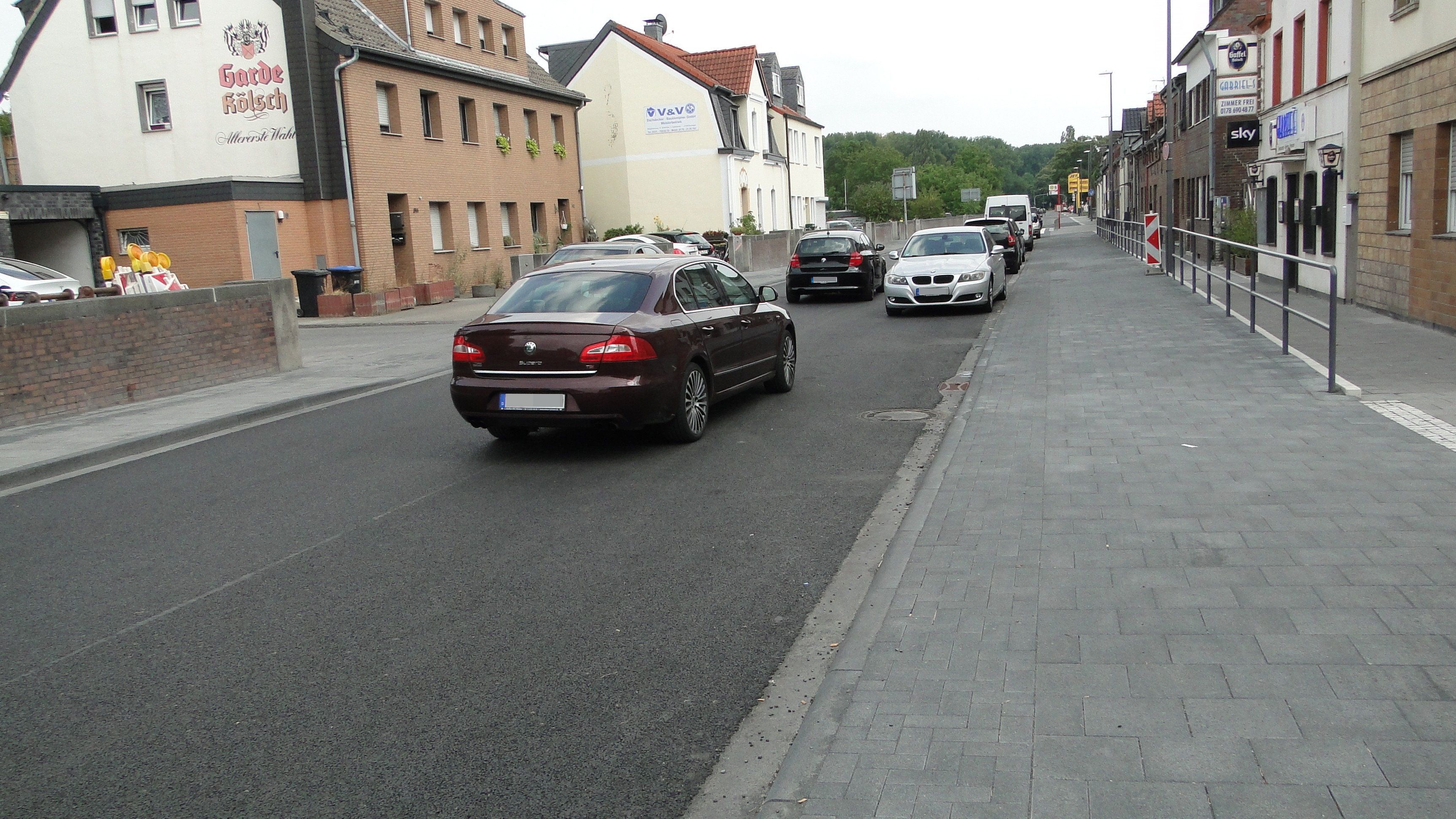 Autos fahren auf einer frisch geteerten Fahrbahn, am Rand parken auch welche.