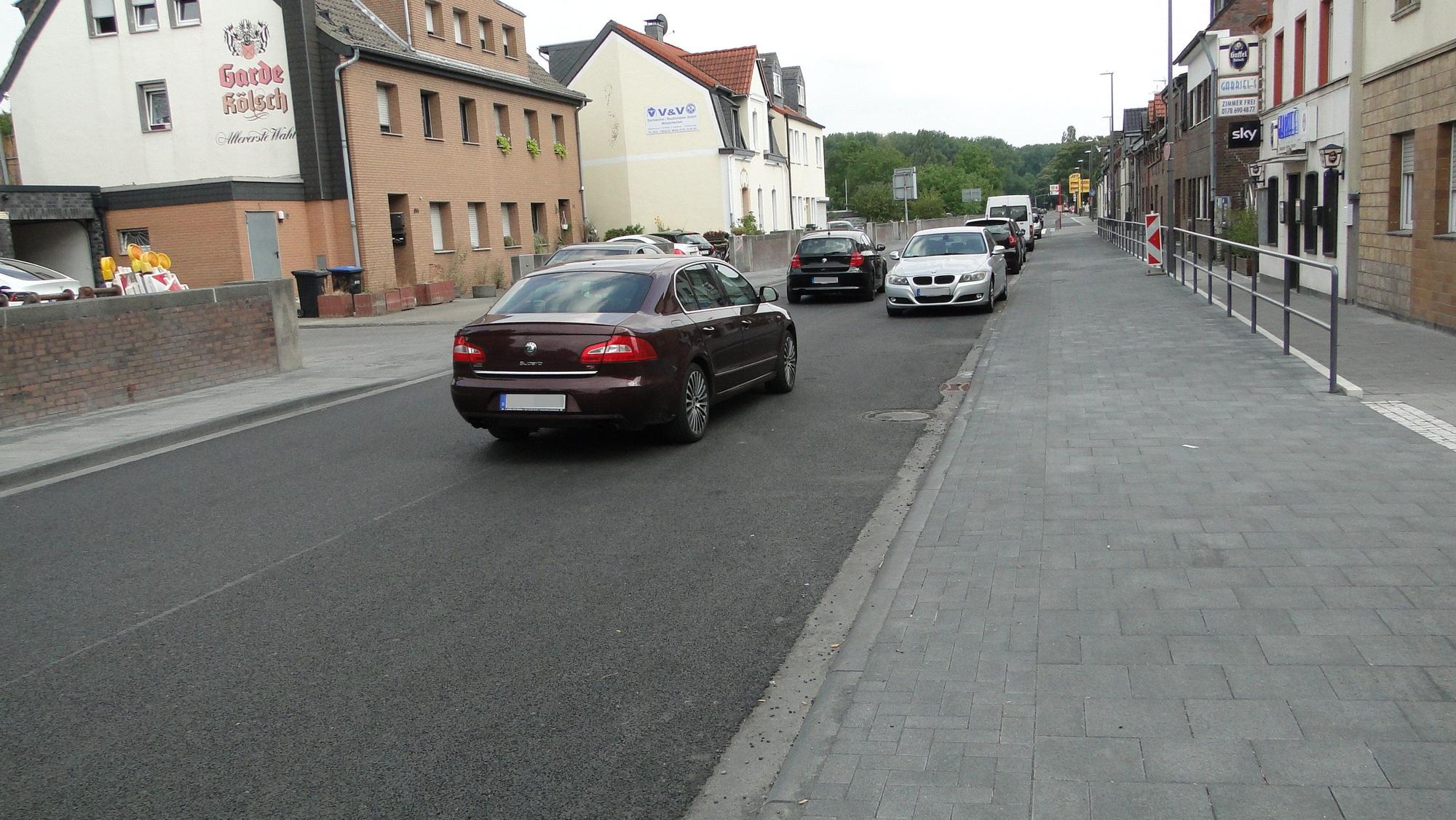Autos fahren auf einer frisch geteerten Fahrbahn, am Rand parken auch welche.