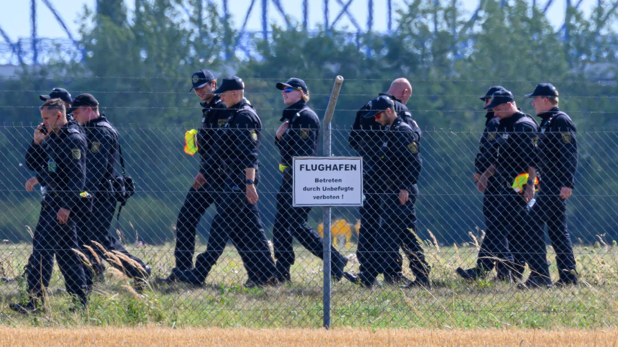 Einsatzkräfte laufen über eine Wiese, davor steht ein Hinweisschild „Flughafen“.