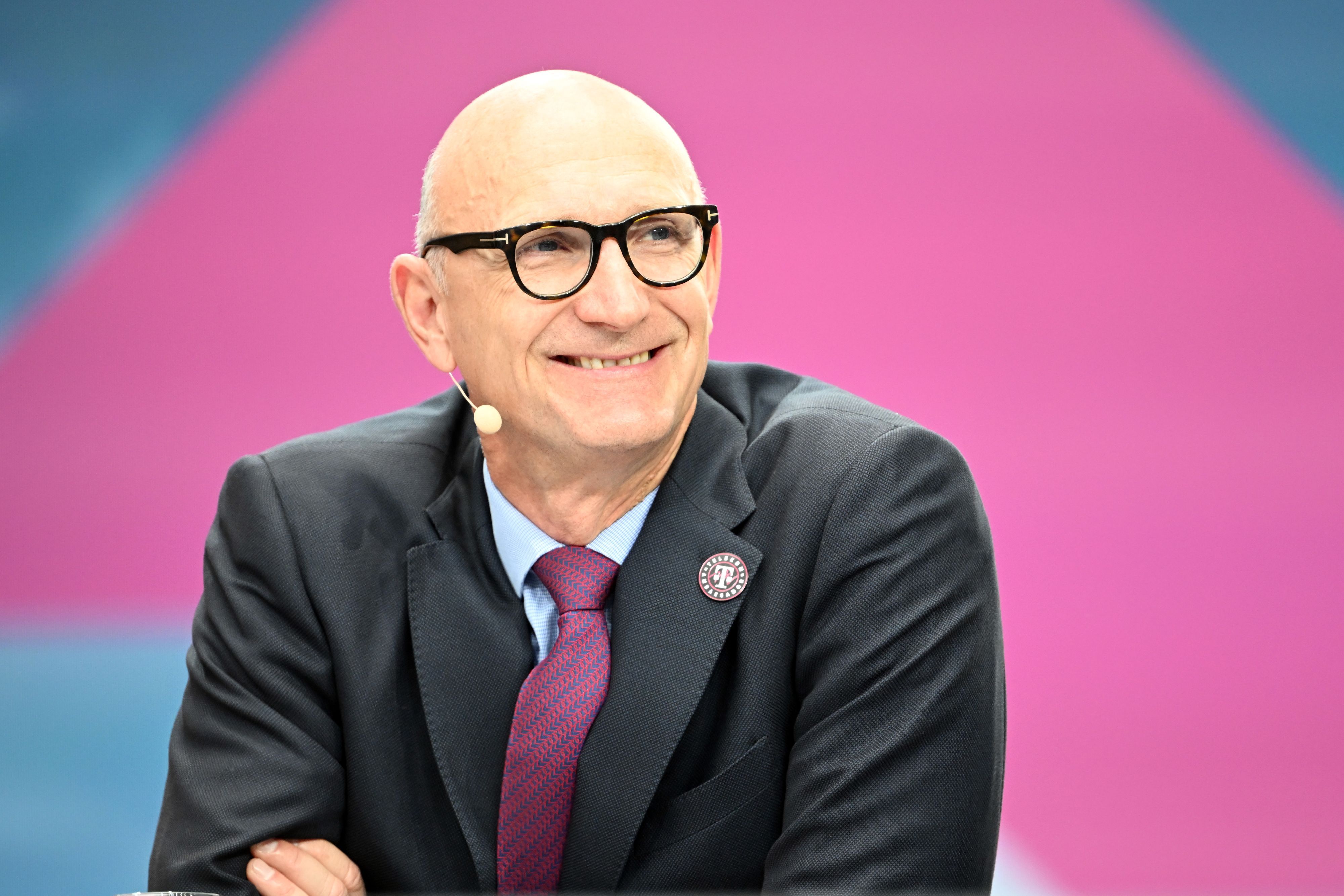 Tim Höttges sitzt vor einem pinken Hintergrund im Anzug und blickt lächelnd an der Kamera vorbei.