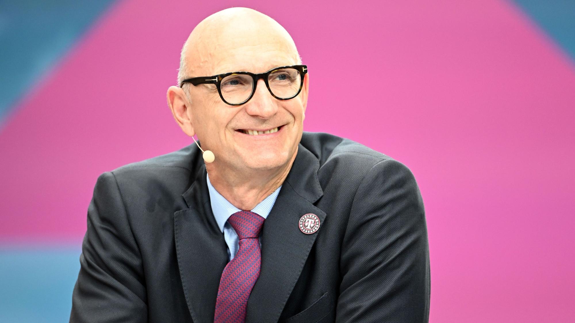 Tim Höttges sitzt vor einem pinken Hintergrund im Anzug und blickt lächelnd an der Kamera vorbei.