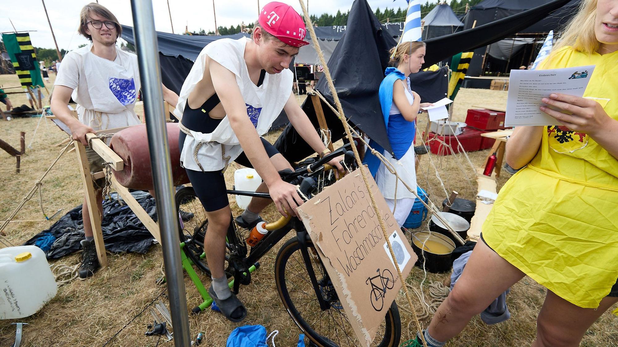 Mit einem Fahrrad treibt ein Jugendlicher, der eine Fahrradkappe trägt, eine Waschtrommel an.