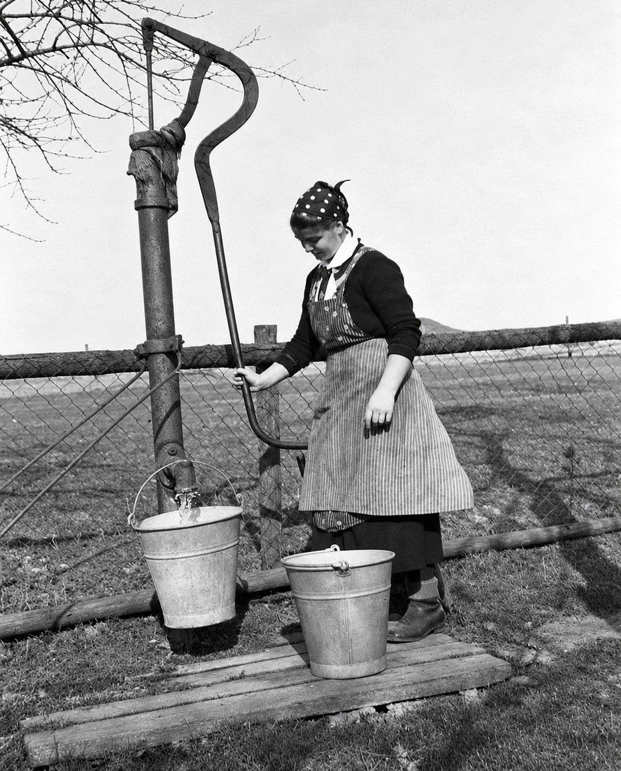 Eine Frau schöpft in den 1930er Jahren in Morsbach mit einer Schwengelpumpe Wasser aus einem Brunnen in zwei Zinkeimer.