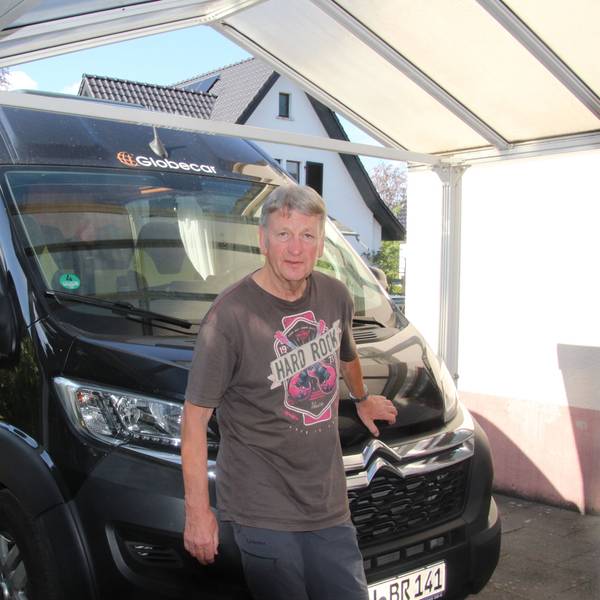 Ein Mann in einem bedruckten T-Shirt steht unter einem Carport vor einem Wohnmobil.
