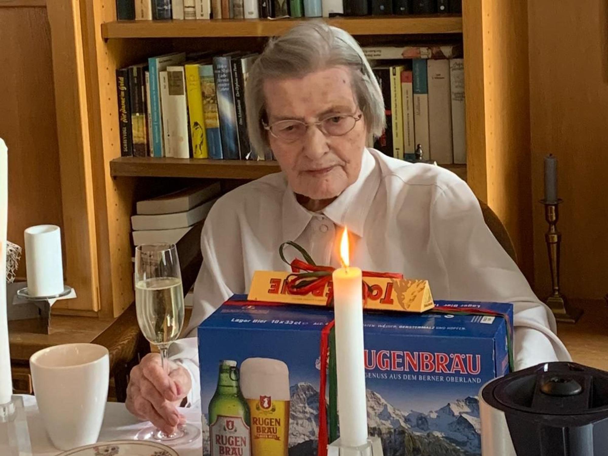 Das Foto zeigt eine ältere Frau mit einem Sektglas in der Hand, vor ihr eine Kerze und eine Packung Bier als Geschenk verpackt.