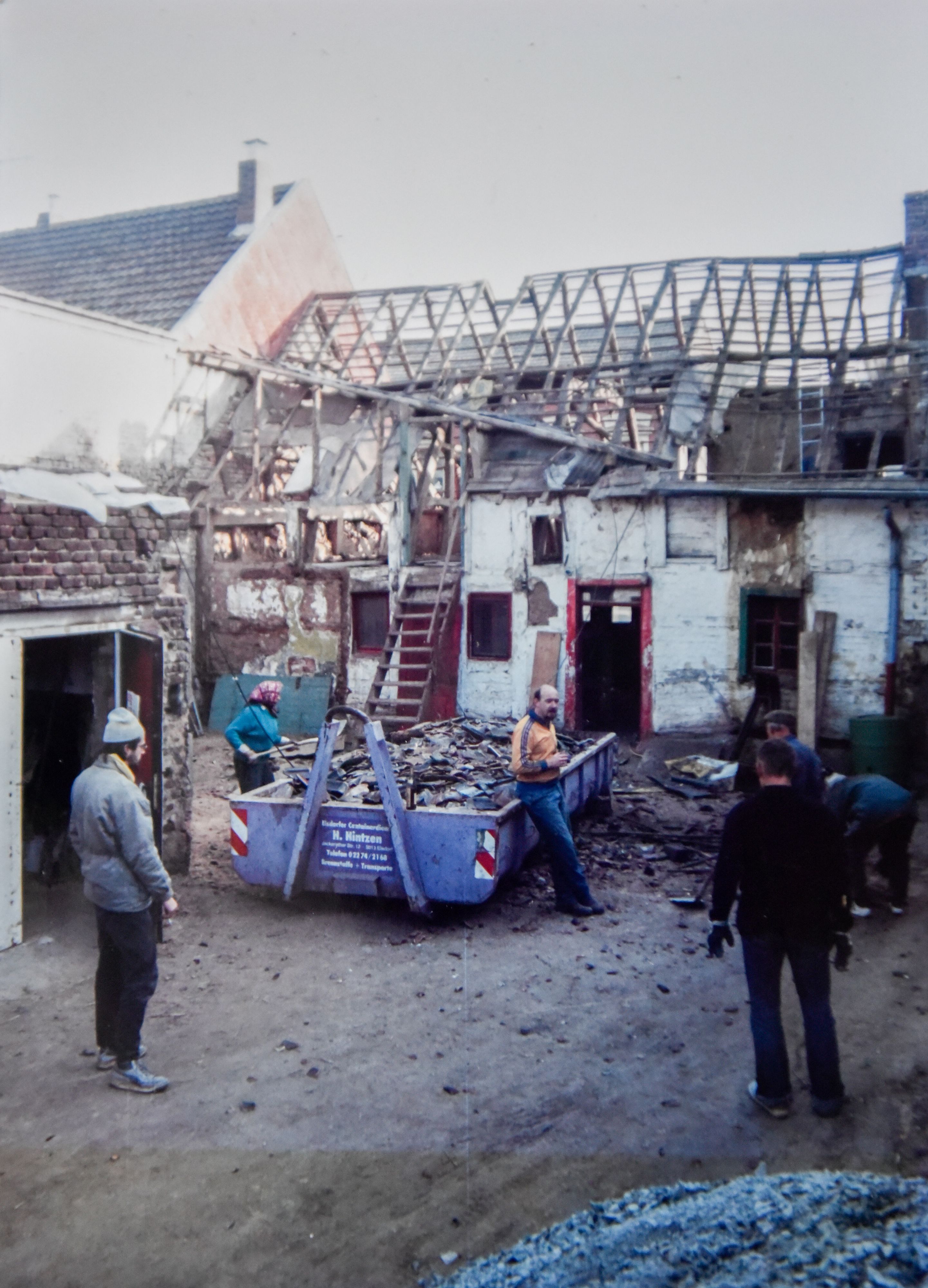 Mehrere Menschen und ein Baucontainer stehen vor einem alten Hof, der verfallen wirkt und restauriert wird.