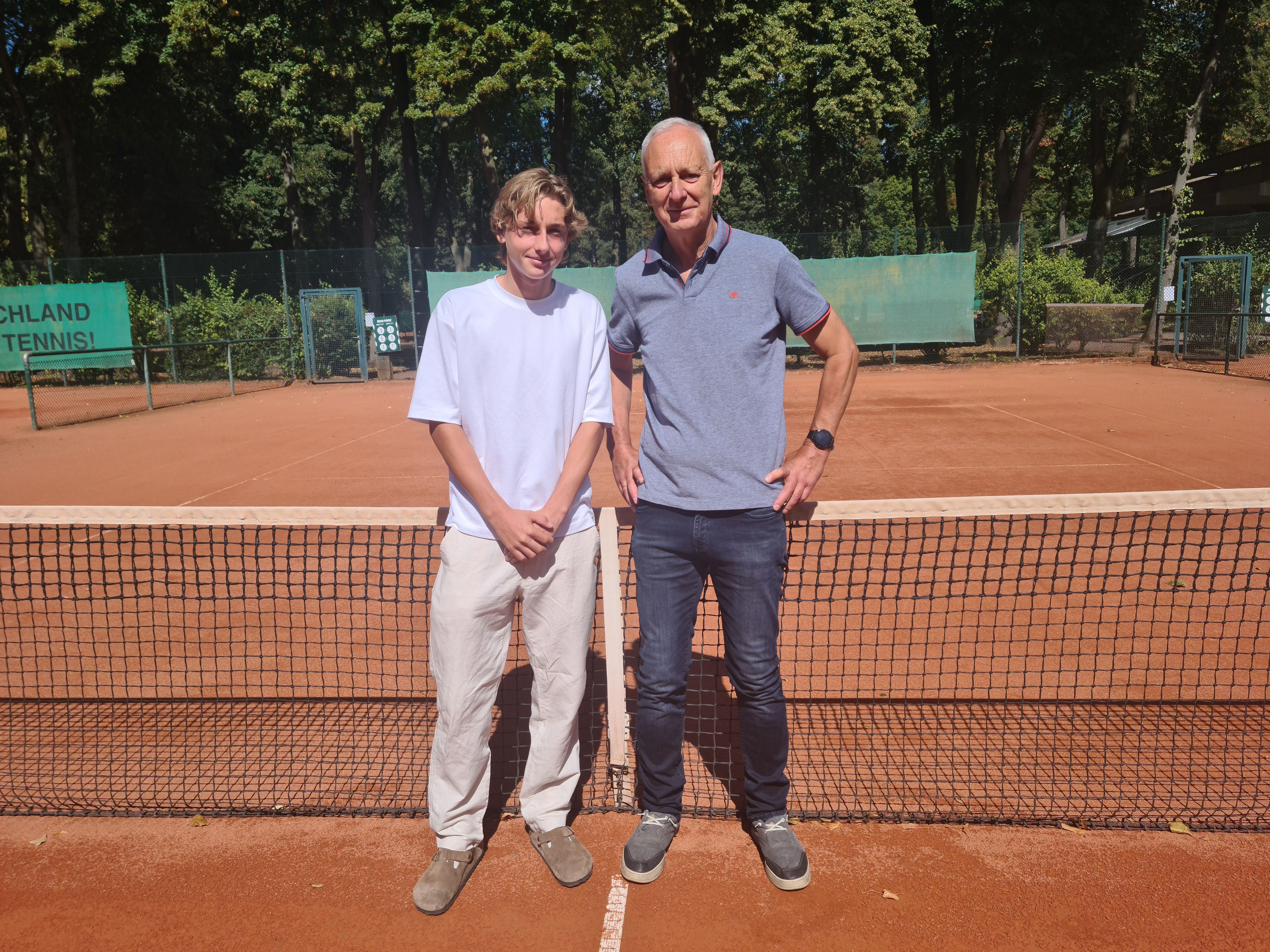 Ein junger Mann im weißen T-Shirt und beigefarbener Hose und ein älterer Mann in Jeans und blauem Polo-Shirt stehen nebeneinander vor einem Tennisnetz auf einem Tennisplatz.