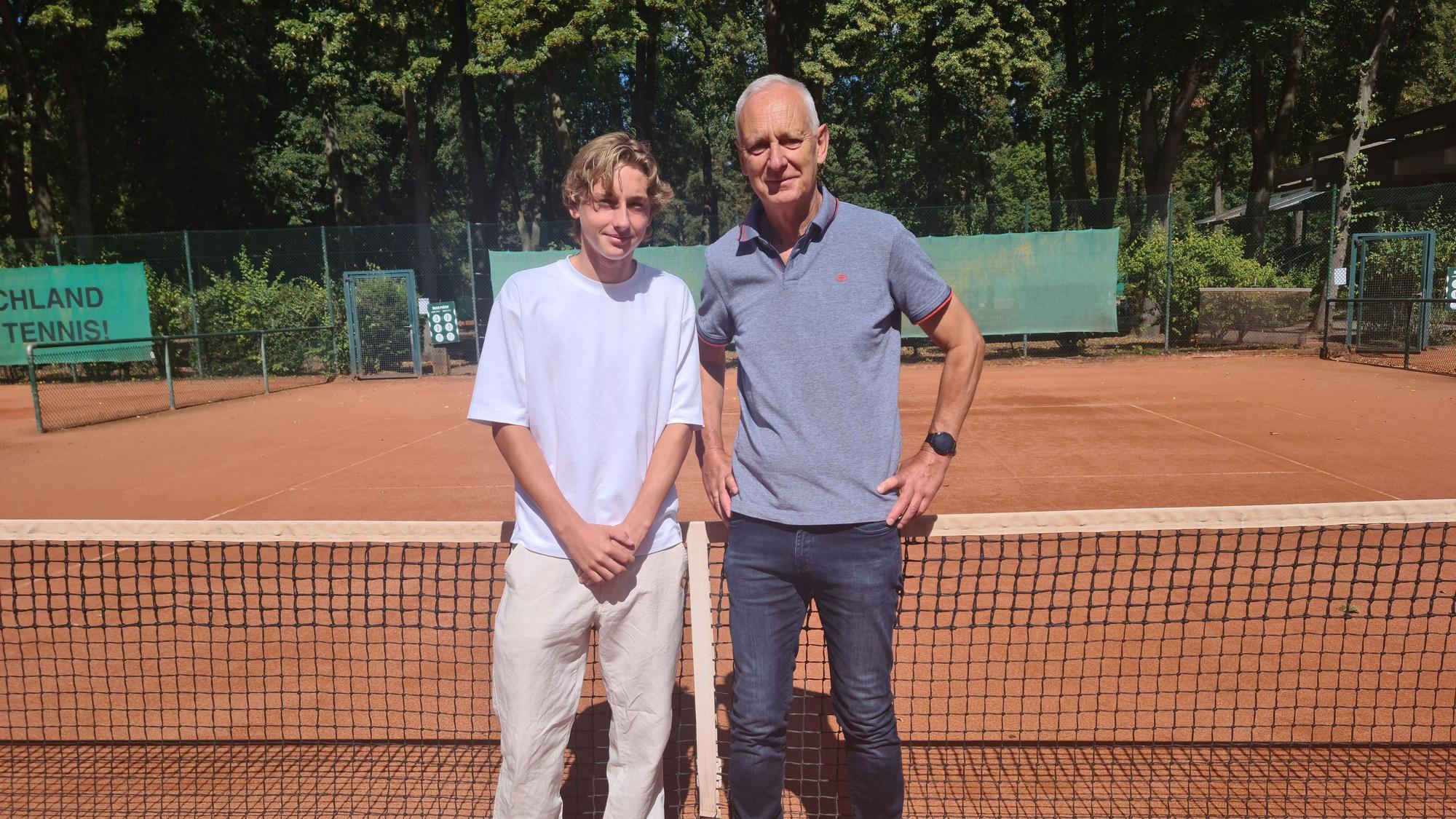 Ein junger Mann im weißen T-Shirt und beigefarbener Hose und ein älterer Mann in Jeans und blauem Polo-Shirt stehen nebeneinander vor einem Tennisnetz auf einem Tennisplatz.