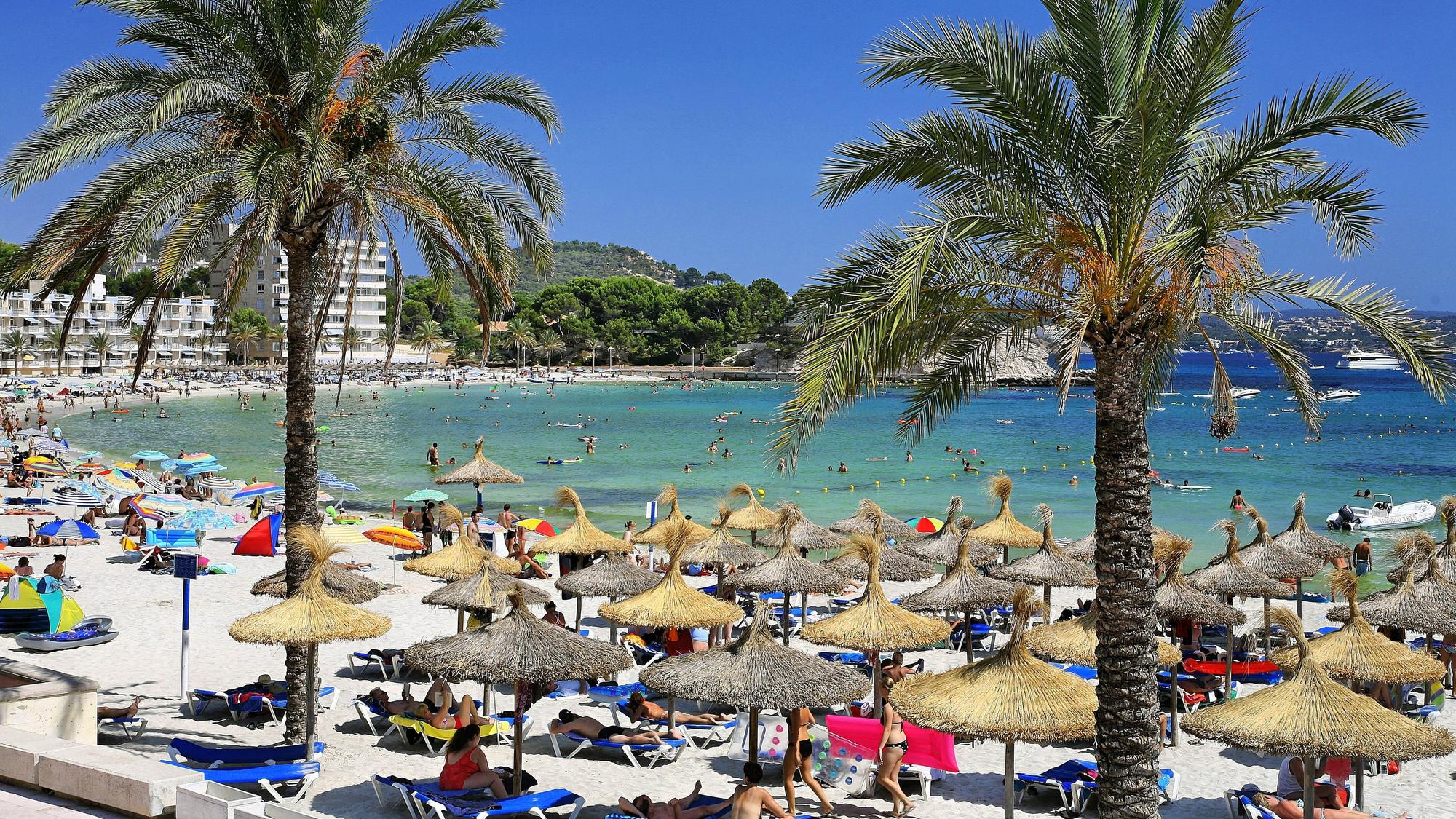 Urlauber an einer Badebucht mit Sonnenschirmen und Palmen.