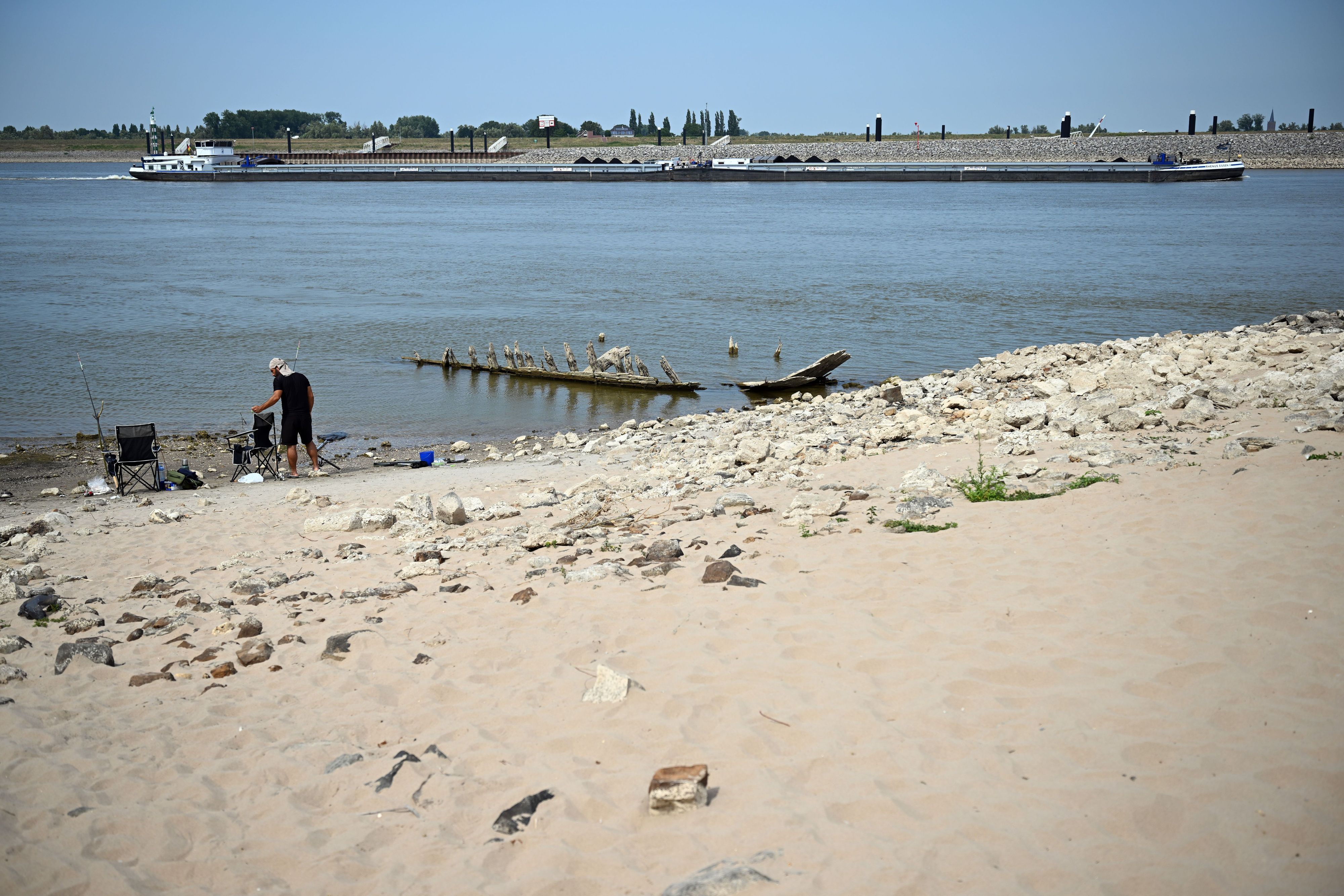 Ein Frachtschiff fährt bei Niedrigwasser auf dem Rhein an dem Wrack eines Bootes vorbei, das normalerweise unter Wasser liegt.