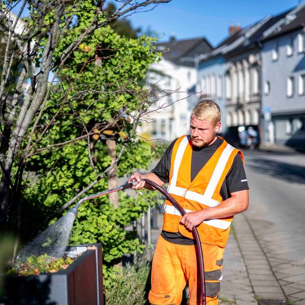 Ein Mann in orange-farbener Kleidung wässert mit einem Schlauch Pflanzen an einem Straßenrand.