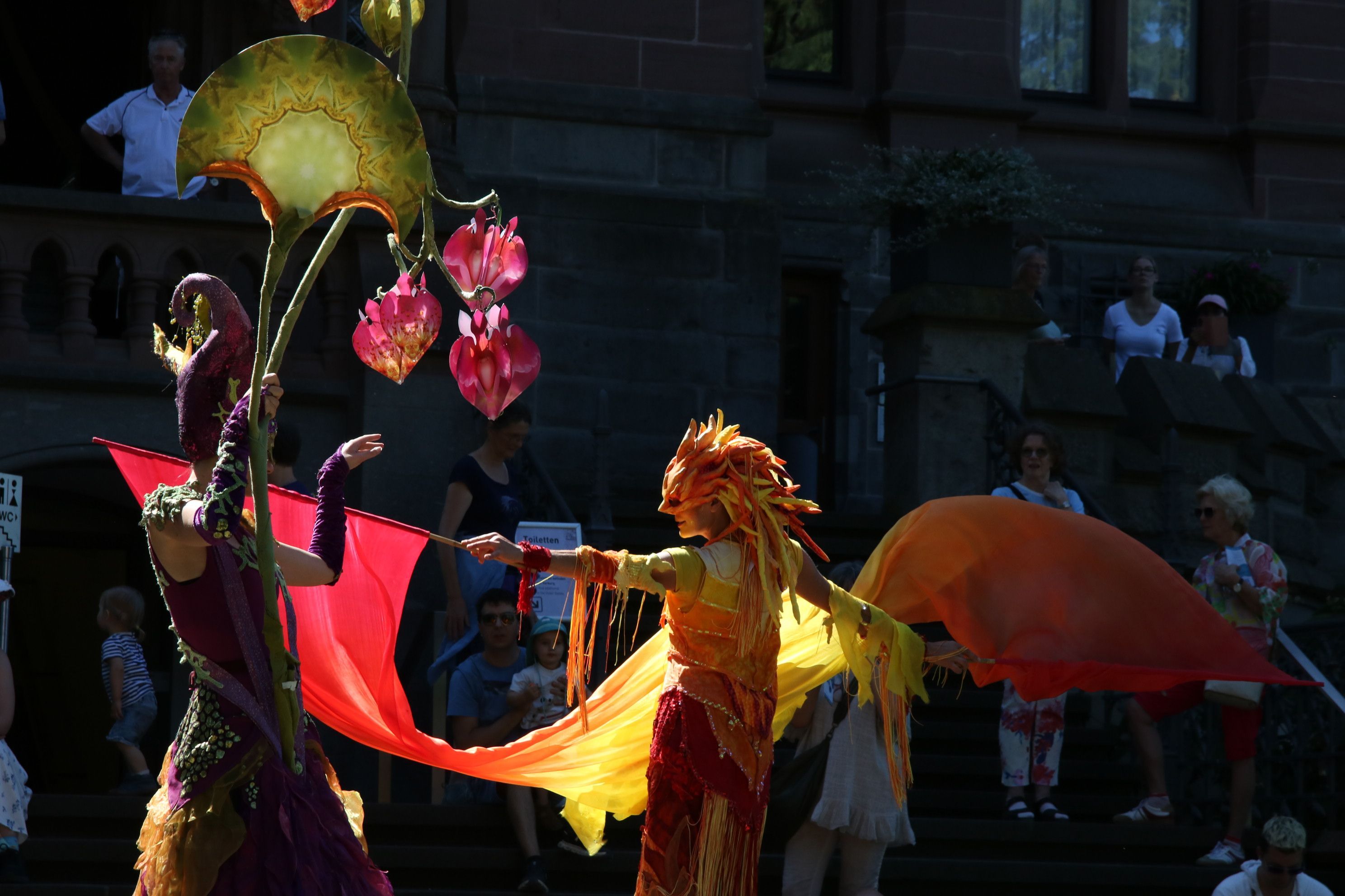 Zwei Darsteller in bunten, im Sonnenlicht gelb und rot leuchtenden Kostümen und mit fantasievollen Masken.