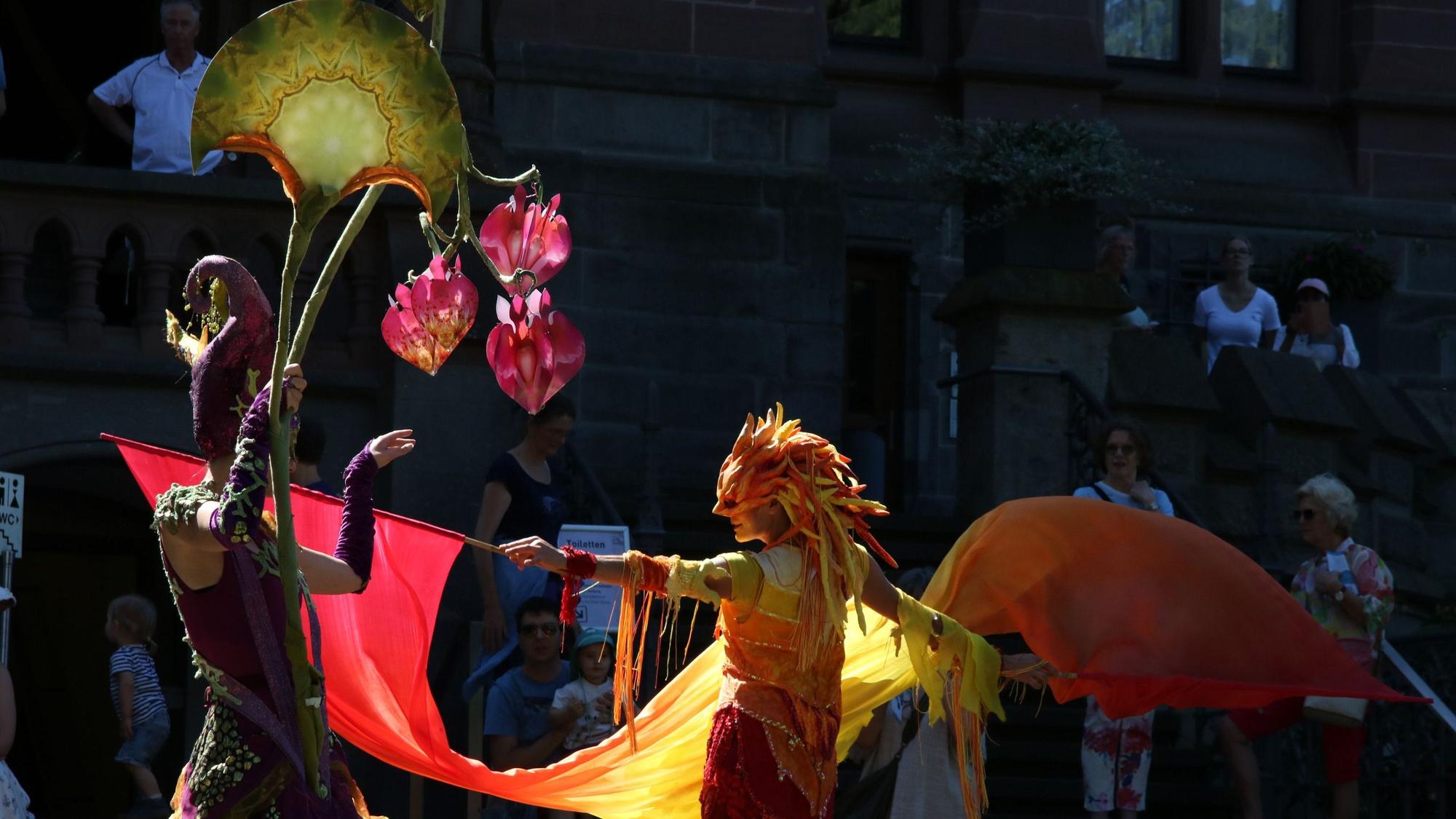 Zwei Darsteller in bunten, im Sonnenlicht gelb und rot leuchtenden Kostümen und mit fantasievollen Masken.