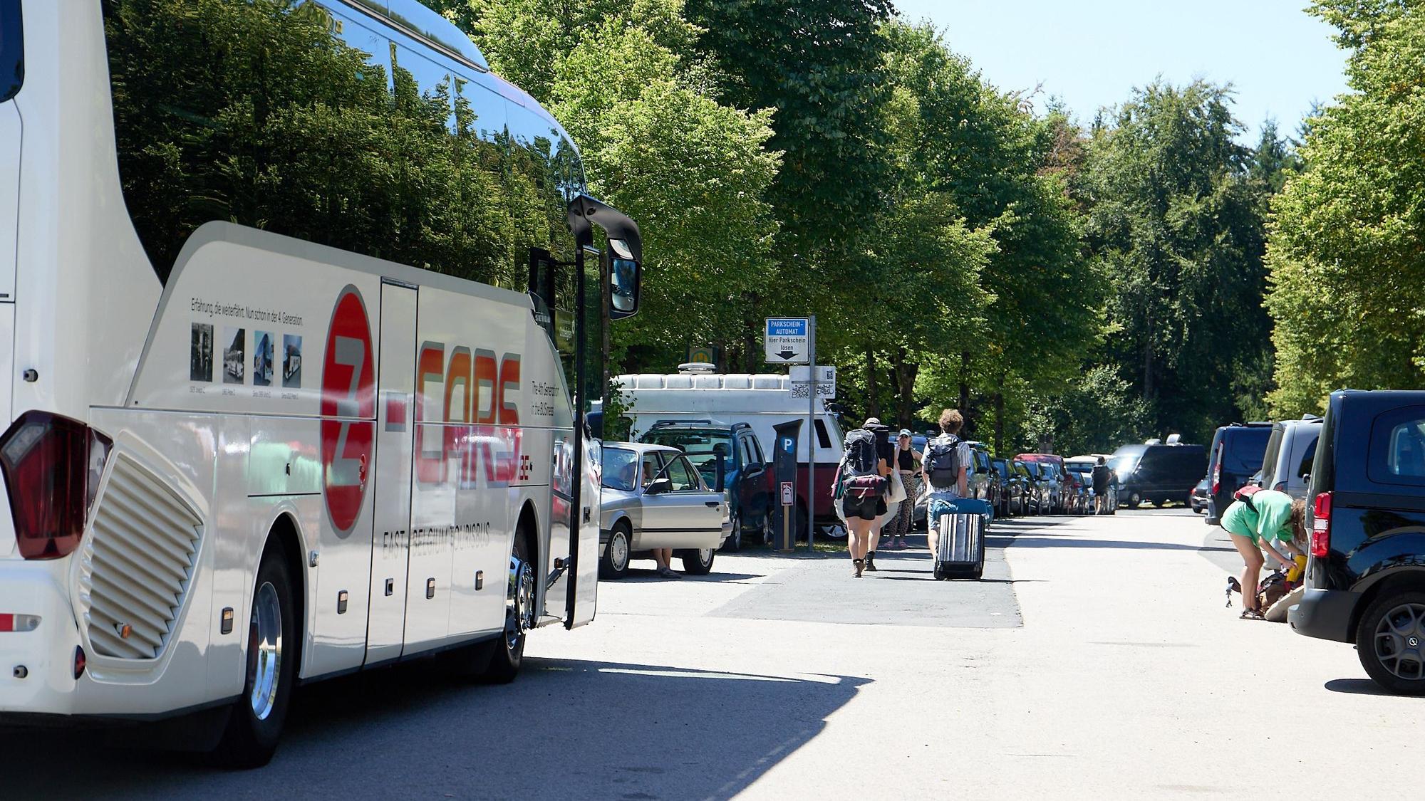 Menschen laufen bepackt über einen Parkplatz, auf dem Busse und Autos parken.