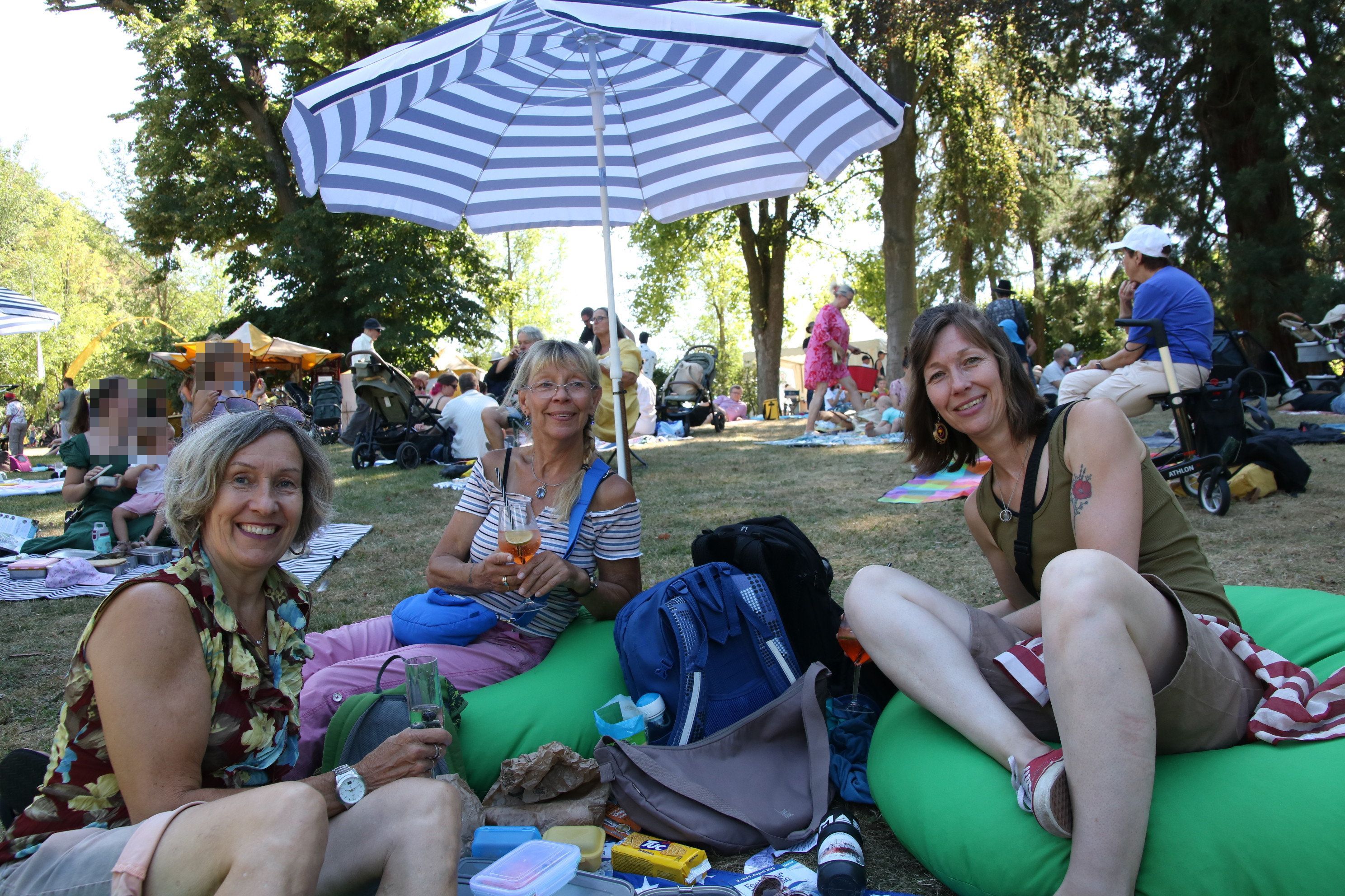 Drei Frauen auf grünen Sitzkissen und unter einem Sonnenschirm im Park.