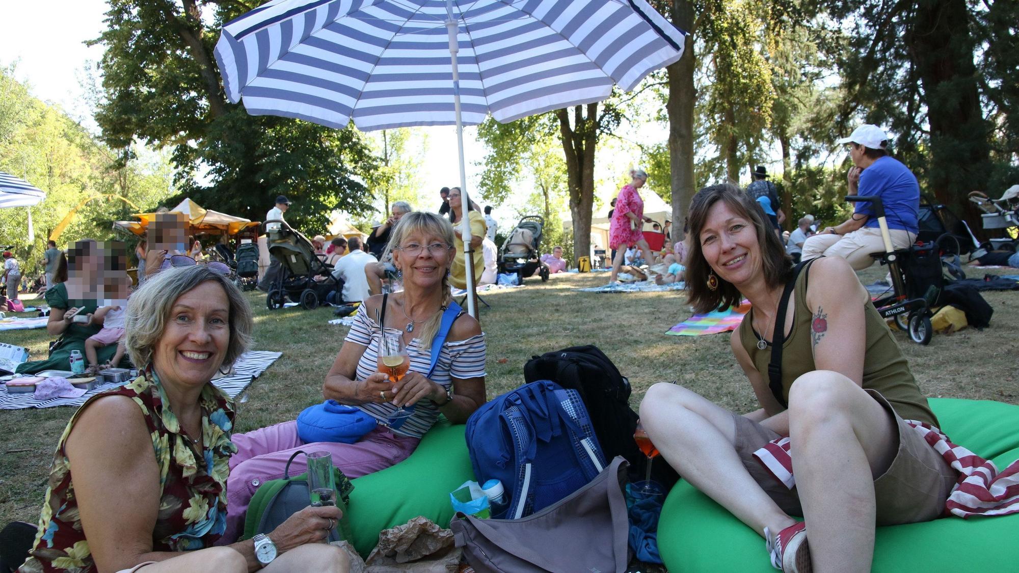 Drei Frauen auf grünen Sitzkissen und unter einem Sonnenschirm im Park.