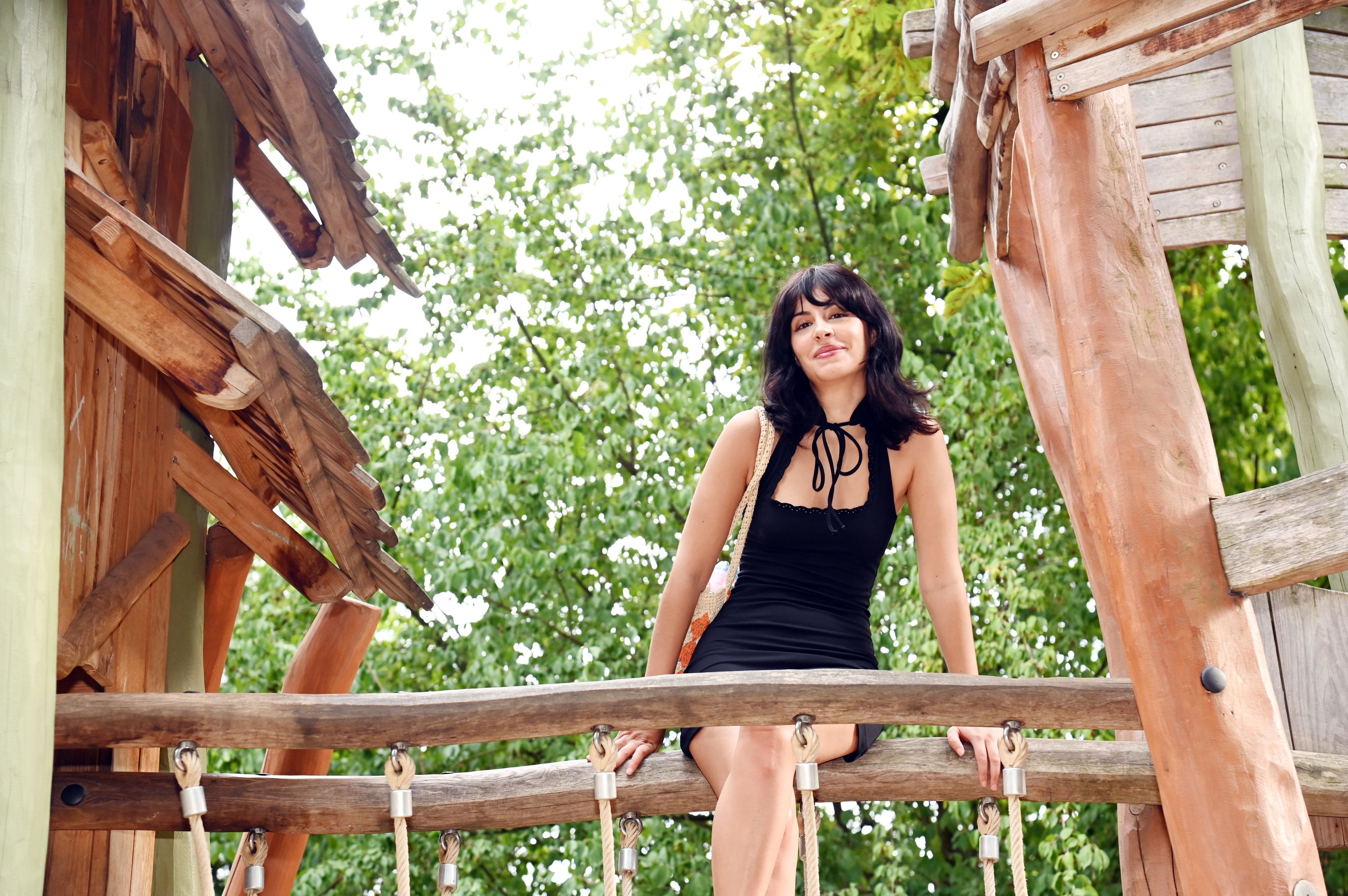 Eine junge Frau mit schwarzem Haar im schwarzen Kleid sitzt auf dem Geländer einer Brücke, die zwei Klettertürme verbindet.