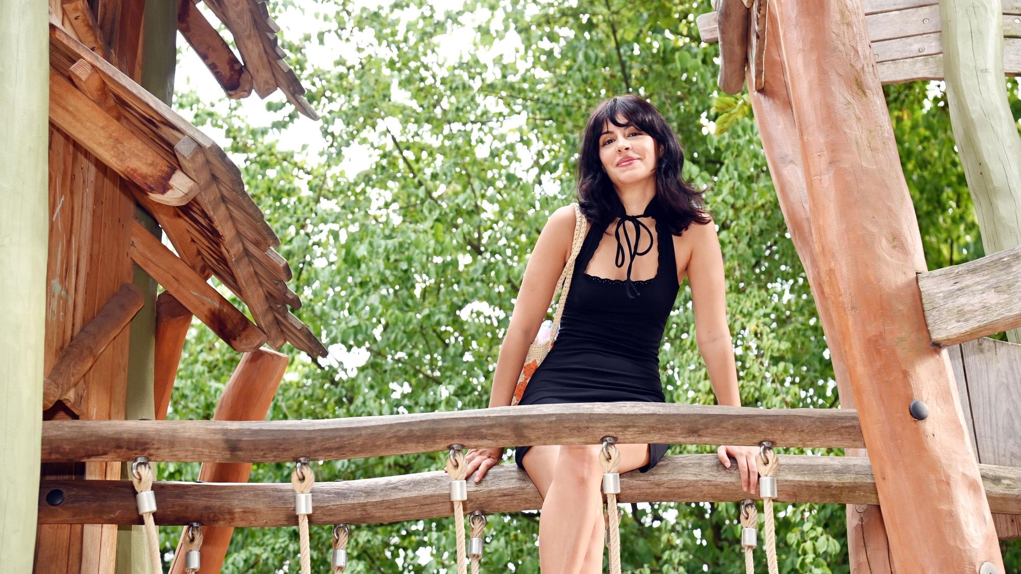 Eine junge Frau mit schwarzem Haar im schwarzen Kleid sitzt auf dem Geländer einer Brücke, die zwei Klettertürme verbindet.