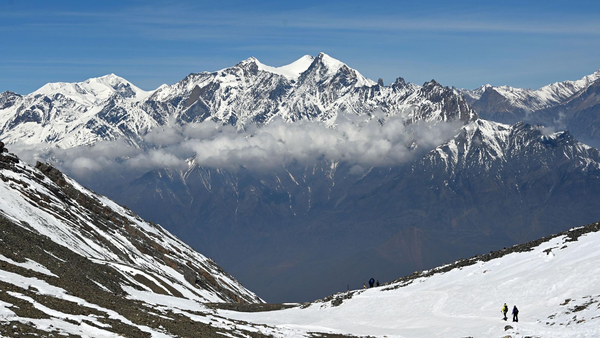 Wanderer gehen am Thorong La Pass in der Annapurna-Region auf einem schneebedeckten Weg, im Hintergrund ragen schneebedeckte Berggipfel des Himalaya über einer Wolkenschicht auf.