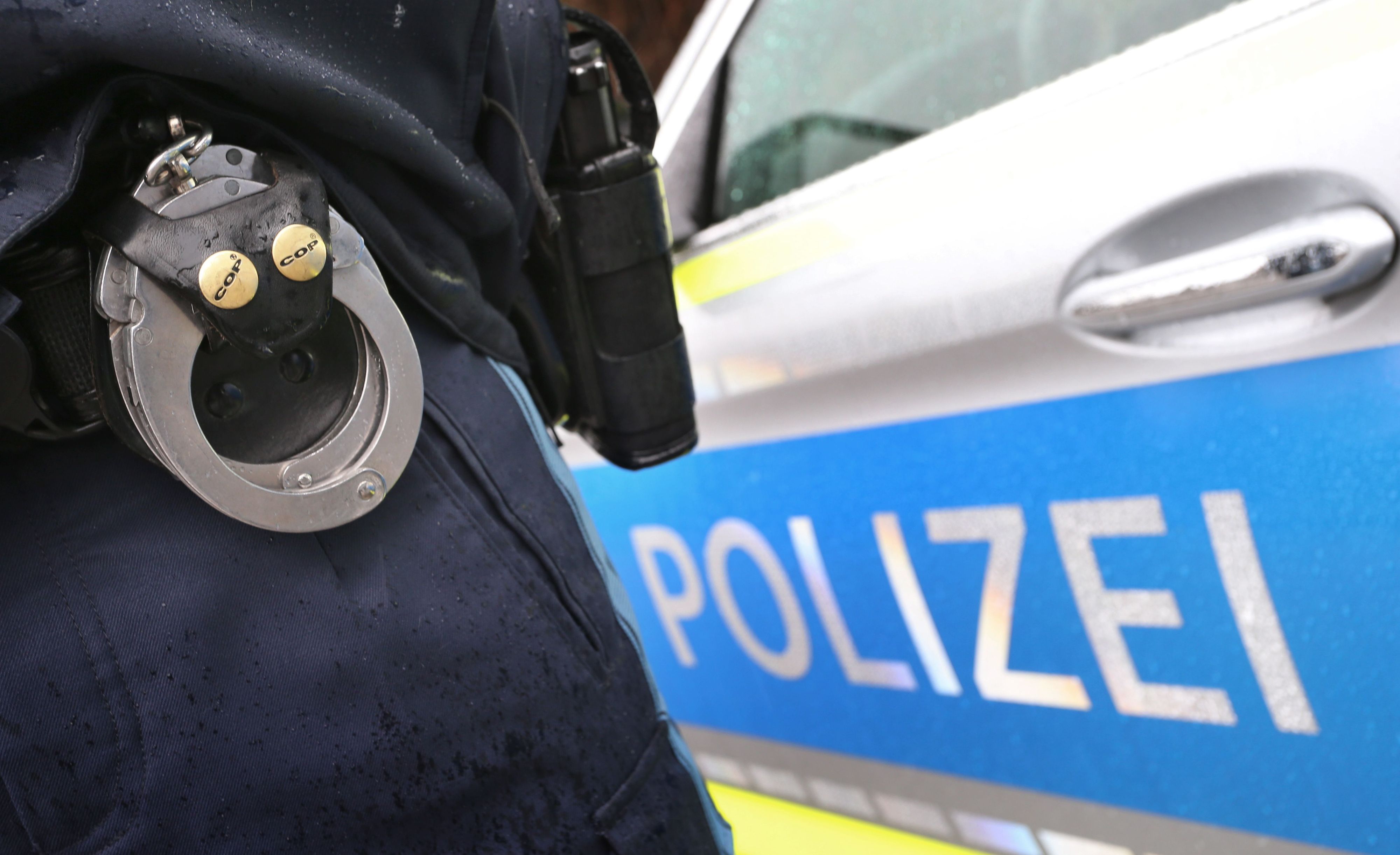 Ein Polizist mit Handschellen und einer Pistole am Gürtel steht vor einem Streifenwagen.