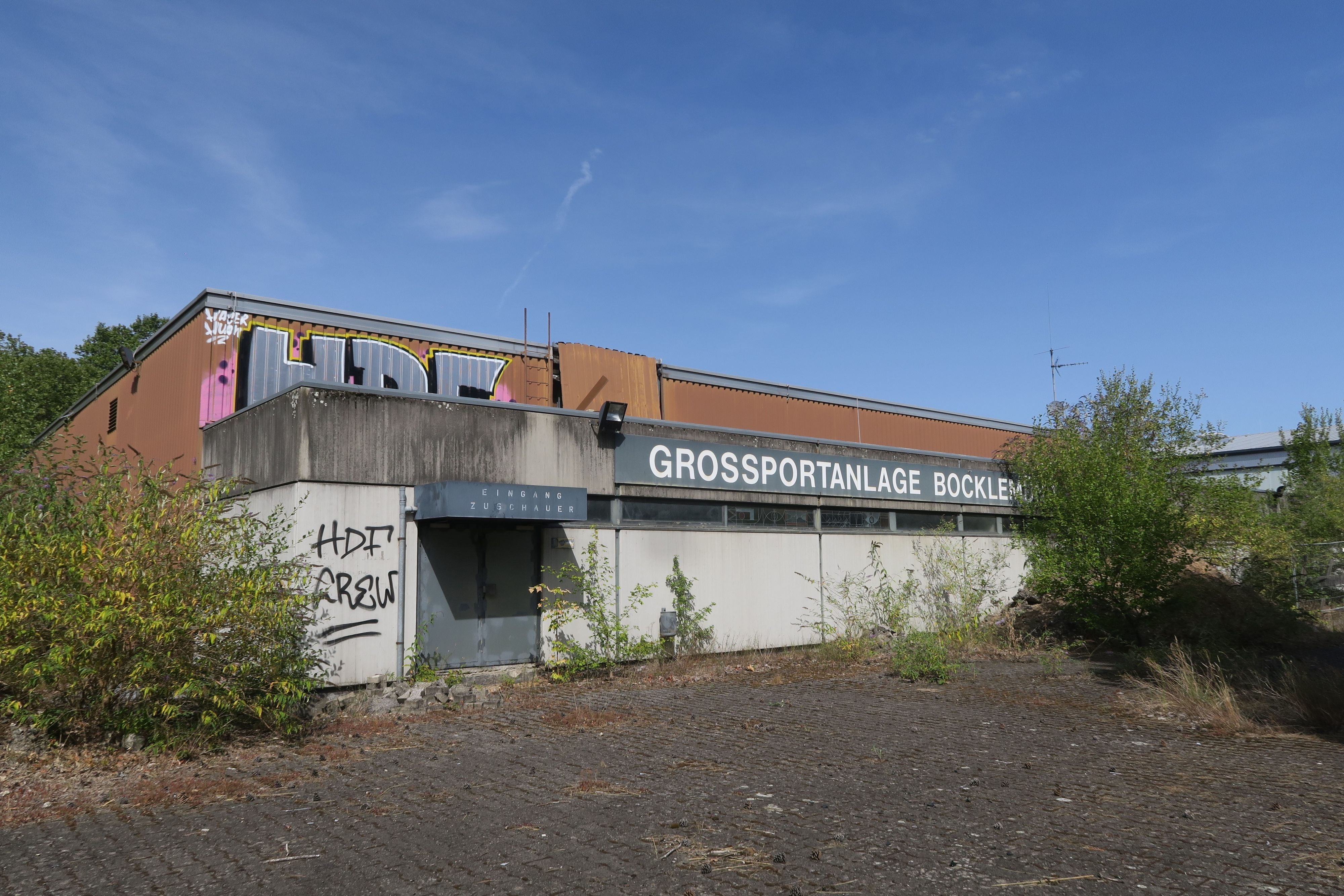 Eine alte Sporthalle, teils mit Graffiti besprüht, trägt den Schriftzug „Großsportanlage Bocklemünd“.