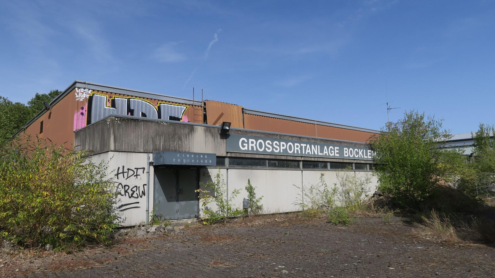 Eine alte Sporthalle, teils mit Graffiti besprüht, trägt den Schriftzug „Großsportanlage Bocklemünd“.