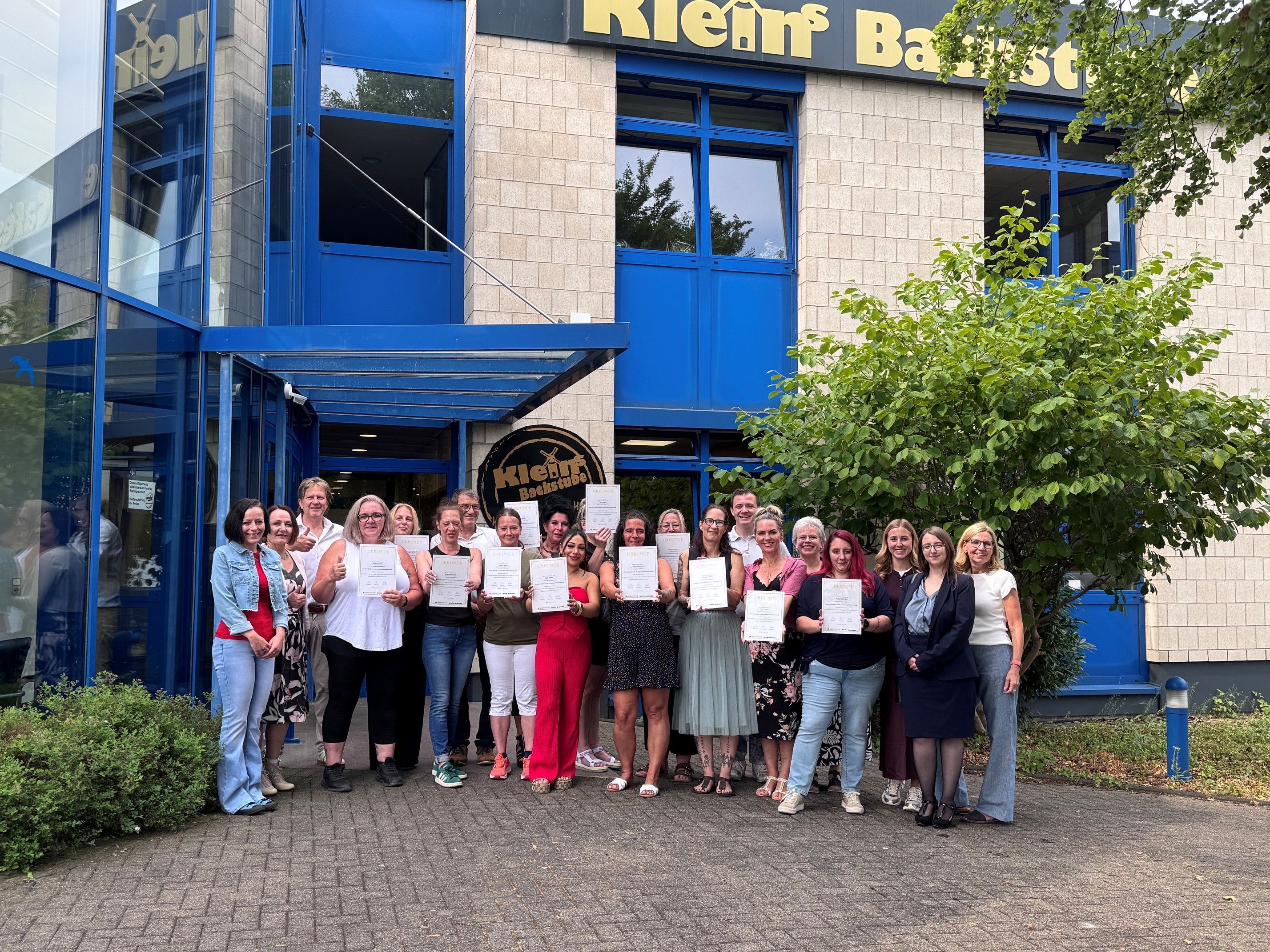 Das Foto zeigt die Absolventinnen mit Urkunden sowie Gratulanten vor der Firmenzentale von „Klein's Backstube“ in Hürth.