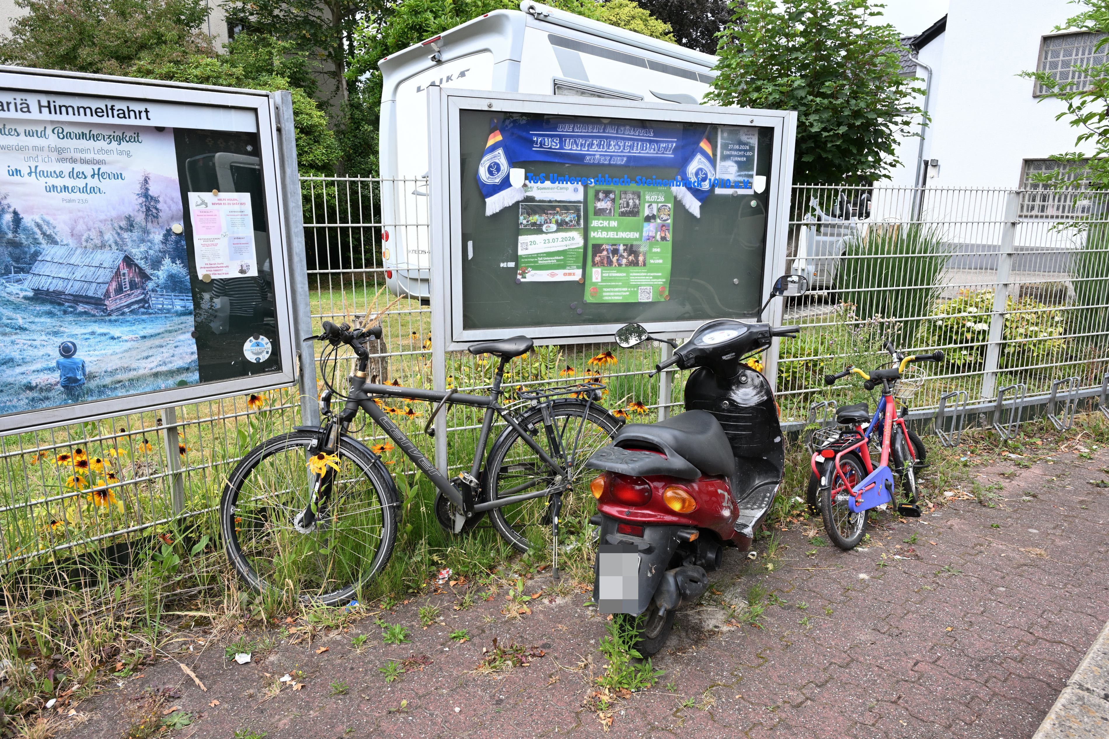 Zu sehen ist ein Fahrrad, zwei Kinderräder und ein Roller, die vor einem Zaun und zwei Infotafeln stehen.